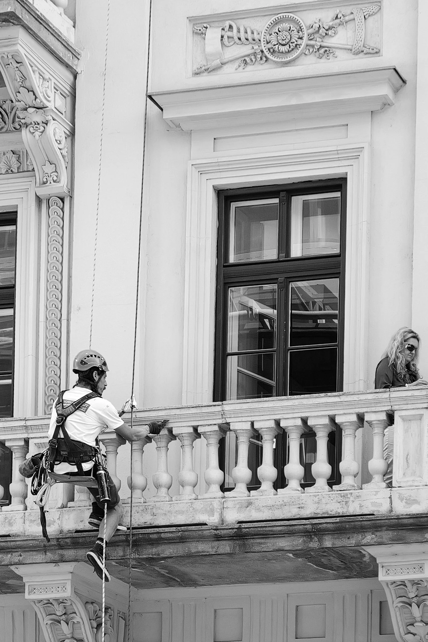 Un homme en casque et équipement de sécurité nettoie la façade d'un bâtiment historique avec des personnages sculptés et des détails architecturaux, tandis qu'une femme avec lunettes regarde par la fenêtre.