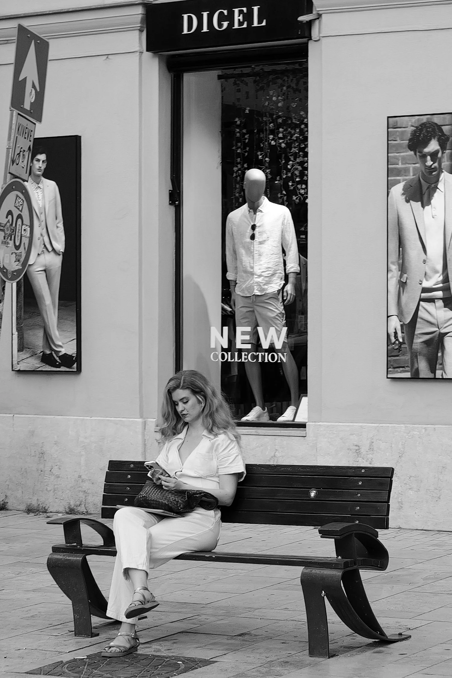 Une femme assise sur un banc en train de regarder son téléphone devant un mur avec deux affiches publicitaires de mode. Au-dessus, une vitrine affiche le texte « NEW COLLECTION » avec un mannequin portant des vêtements décontractés.
