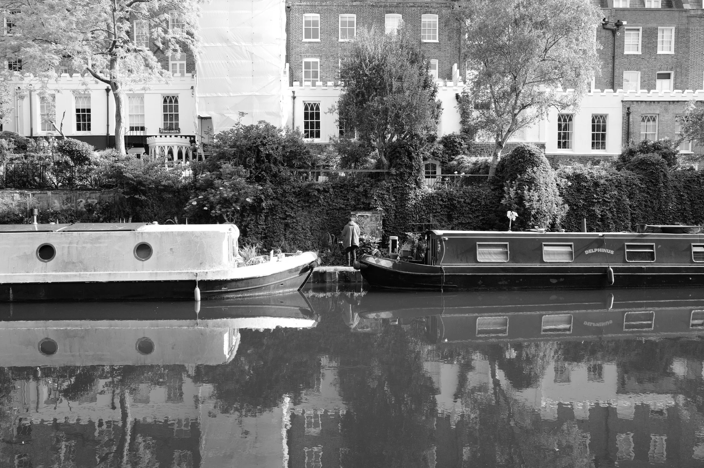 Une scène urbaine avec un canal où sont amarrés deux bateaux, un homme se tenant près de l'eau, des arbres et des bâtiments résidentiels en arrière-plan, en noir et blanc.