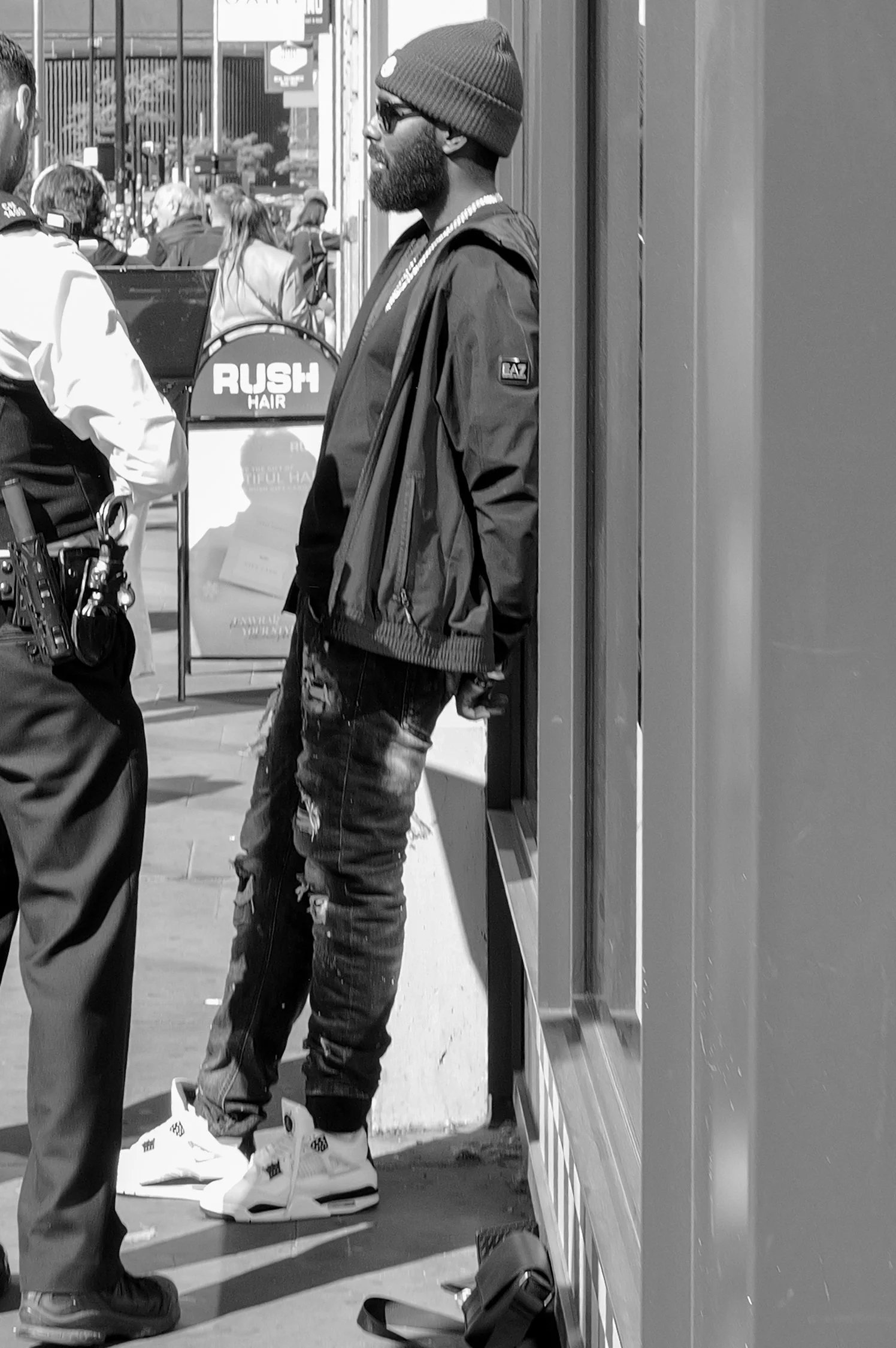 Un homme portant une casquette, des lunettes, un collier, une veste et un jean déchiré, debout à côté d'un mur, dans une rue animée avec des passants et des panneaux publicitaires.