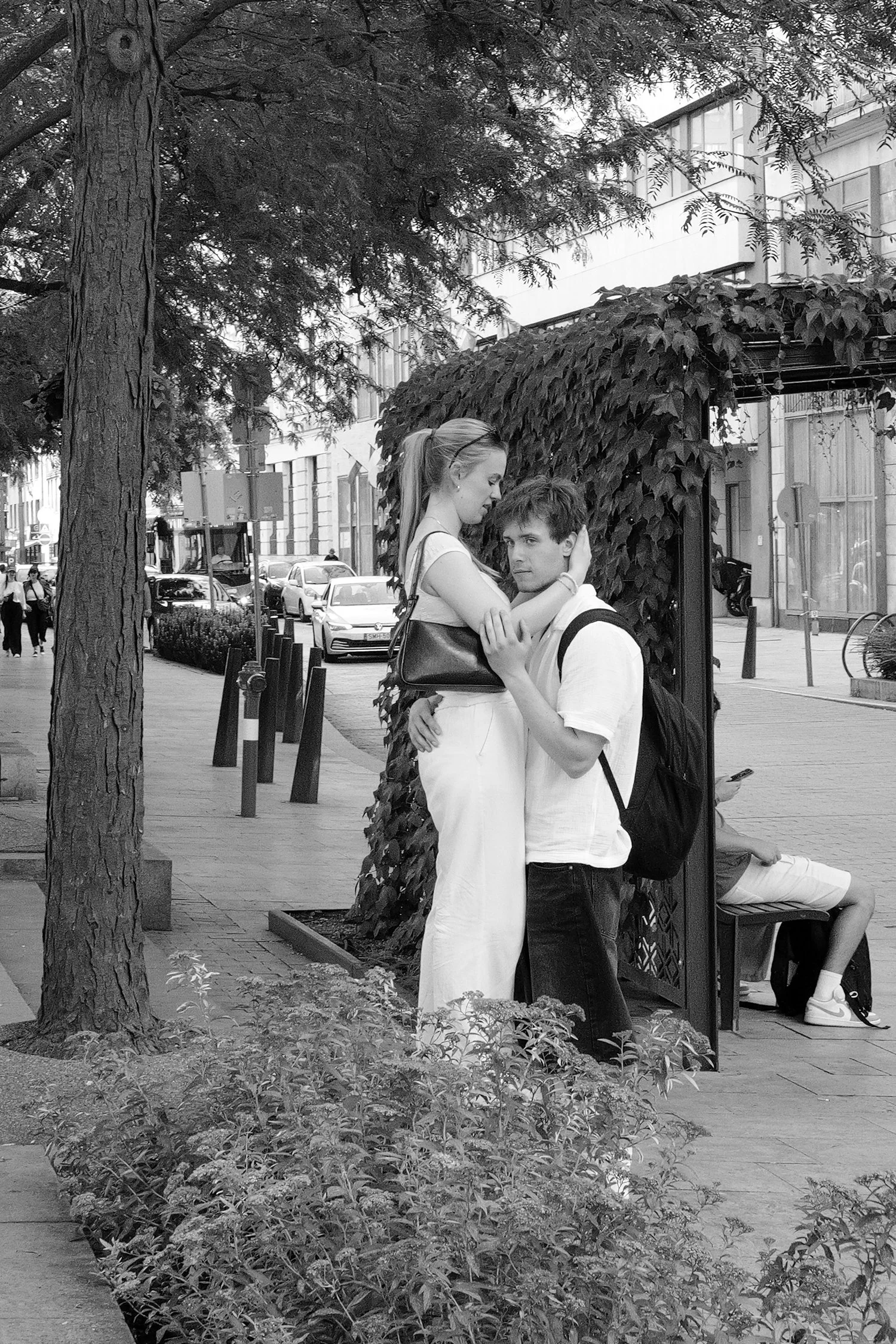 Une jeune femme et un jeune homme s'embrassent dans une rue urbaine, avec des bâtiments modernes, un arbre, une cabine téléphonique et des voitures en arrière-plan, en noir et blanc.