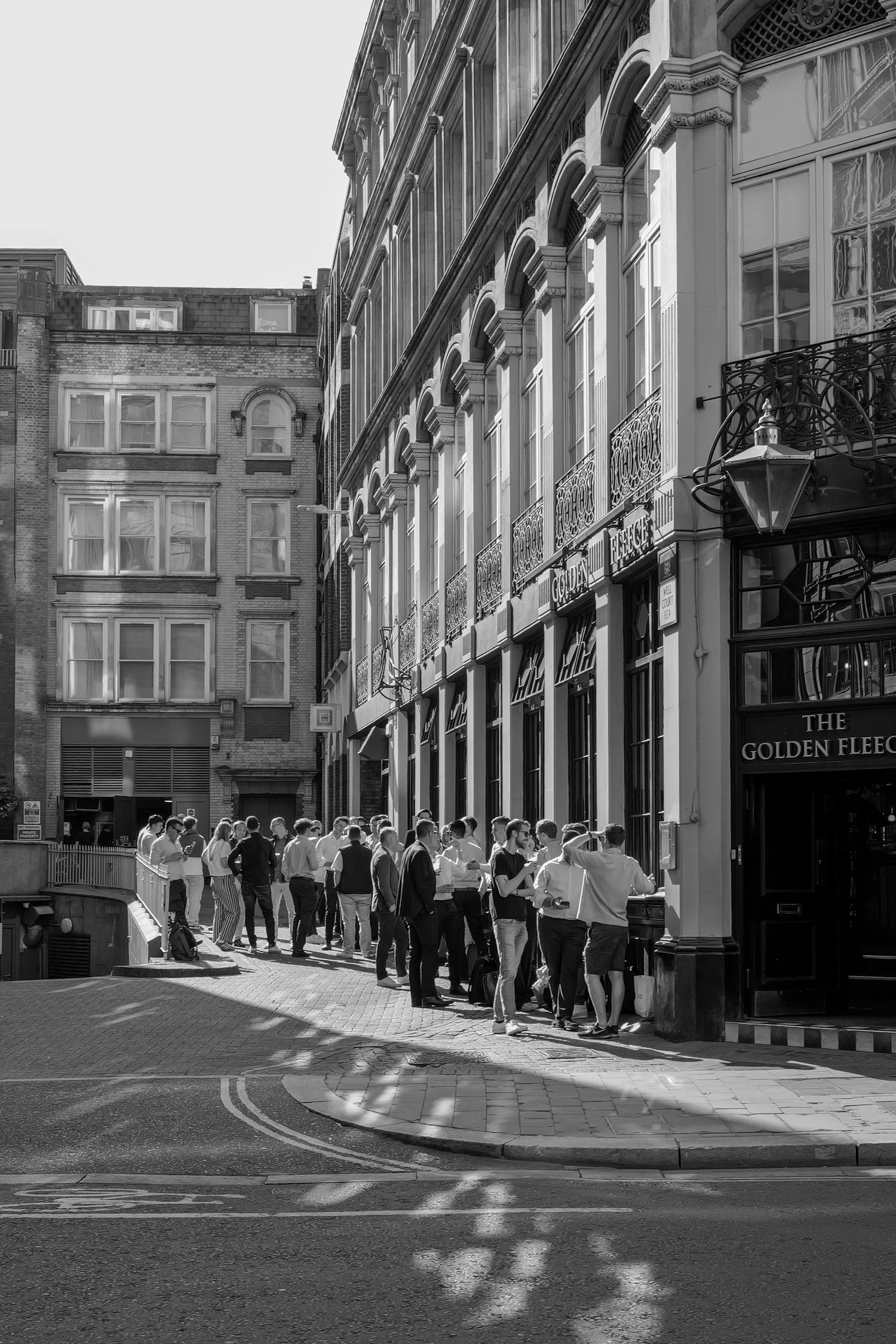 Groupe de personnes faisant la queue devant un pub nommé 'The Golden Fleec'. Les gens se tiennent sous un lampadaire en haut d'une rue pavée bordée de bâtiments anciens. La scène est en noir et blanc.