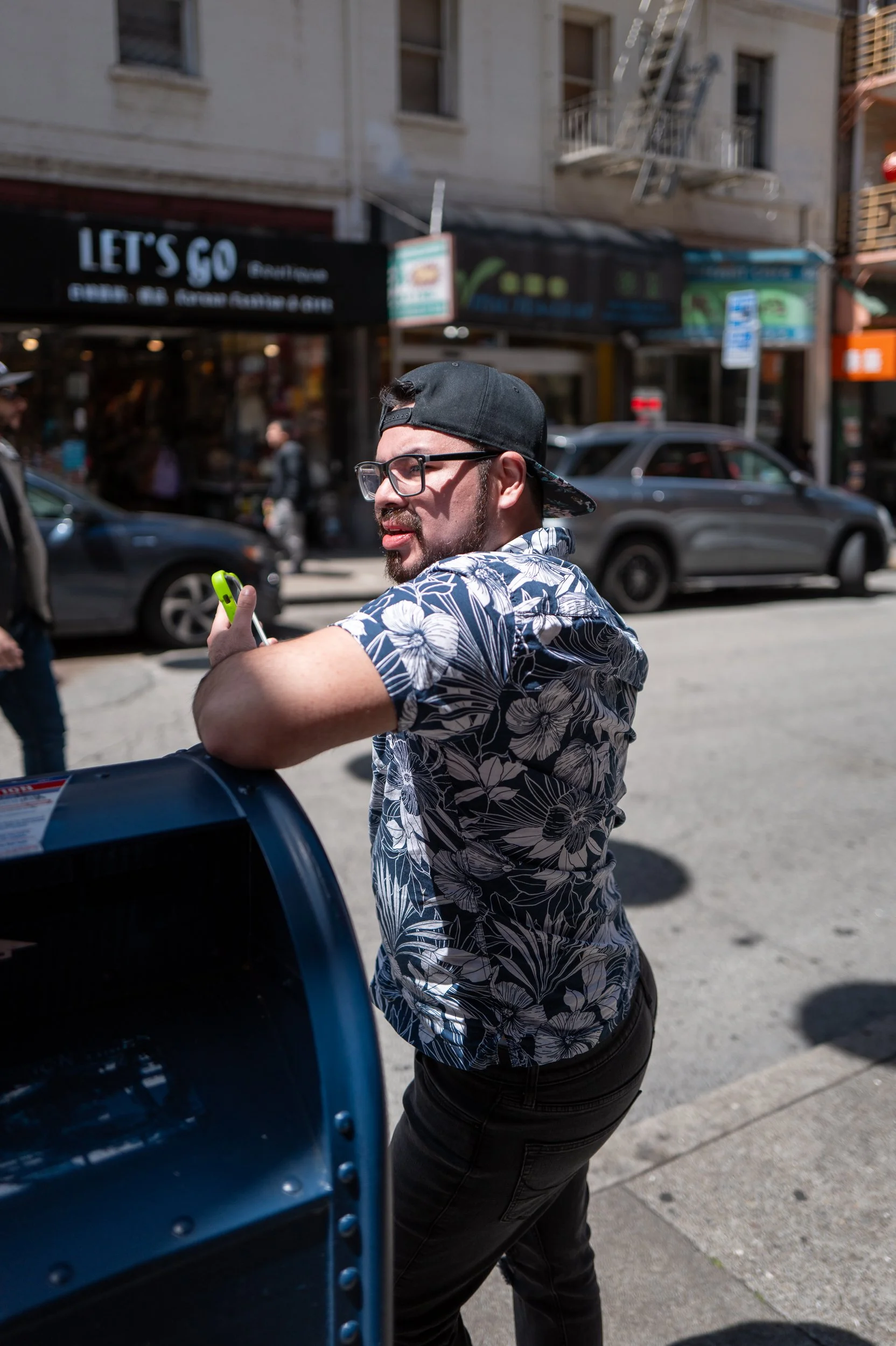 Un homme portant une chemise à motif floral, un bonnet à l'envers, et des lunettes, se tient à côté d'une boîte aux lettres sur le trottoir, en milieu urbain.