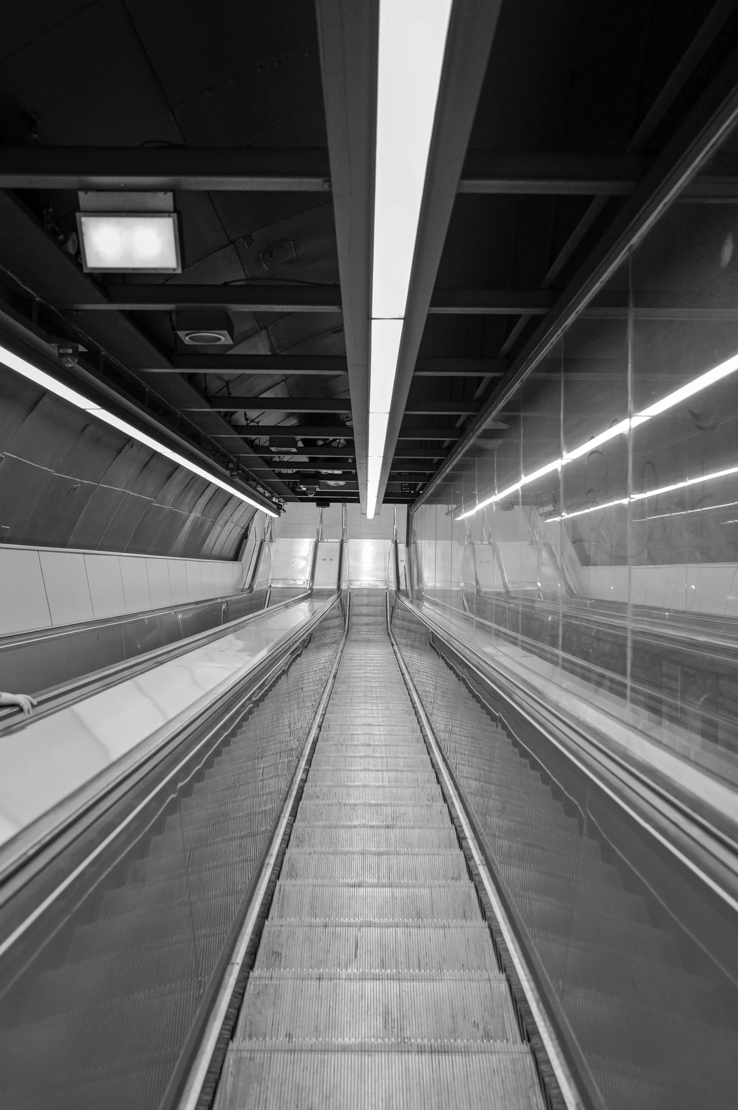 Escalators dans une station de métro ou un centre commercial en noir et blanc, avec des rampes métalliques et un plafond sombre.
