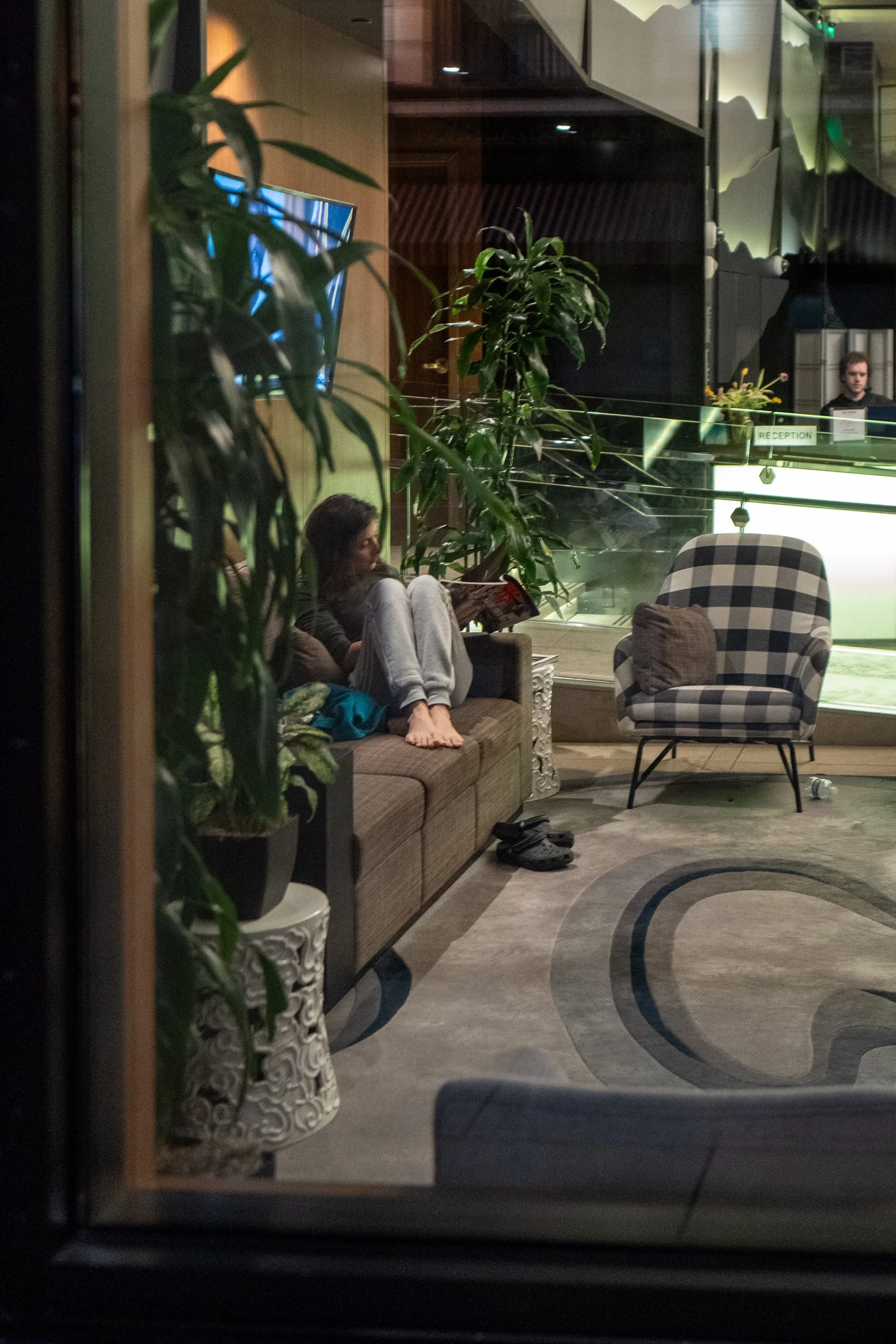 Une femme assise sur un canapé, lisant un magazine dans un hall d'hôtel, avec un fauteuil à côté et des plantes vertes autour, vue depuis une fenêtre, en soirée.