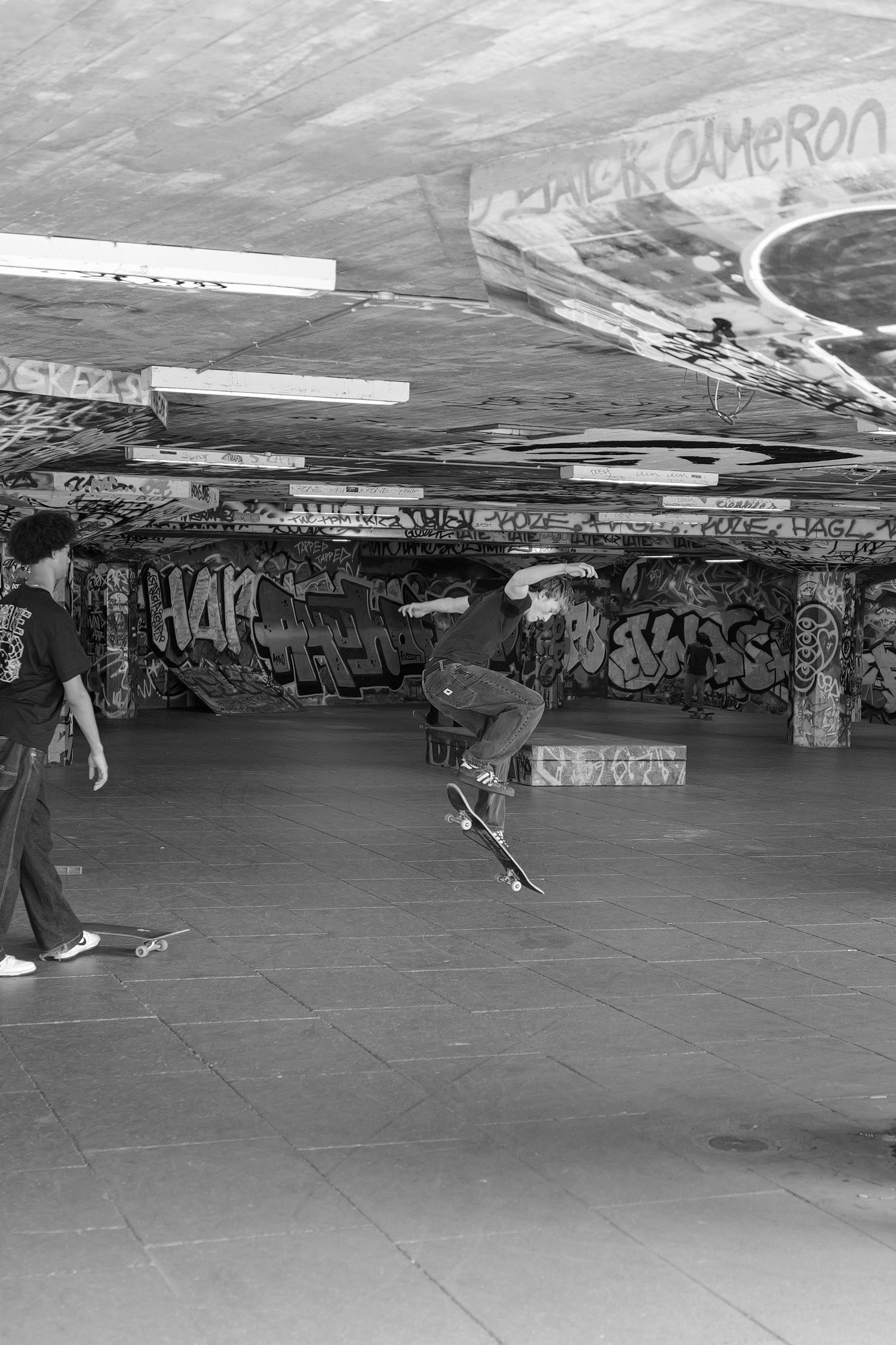 Jeunes pratiquant le skateboard dans un skatepark graffité, sous un plafond recouvert de tags et de graffitis. La scène est en noir et blanc.