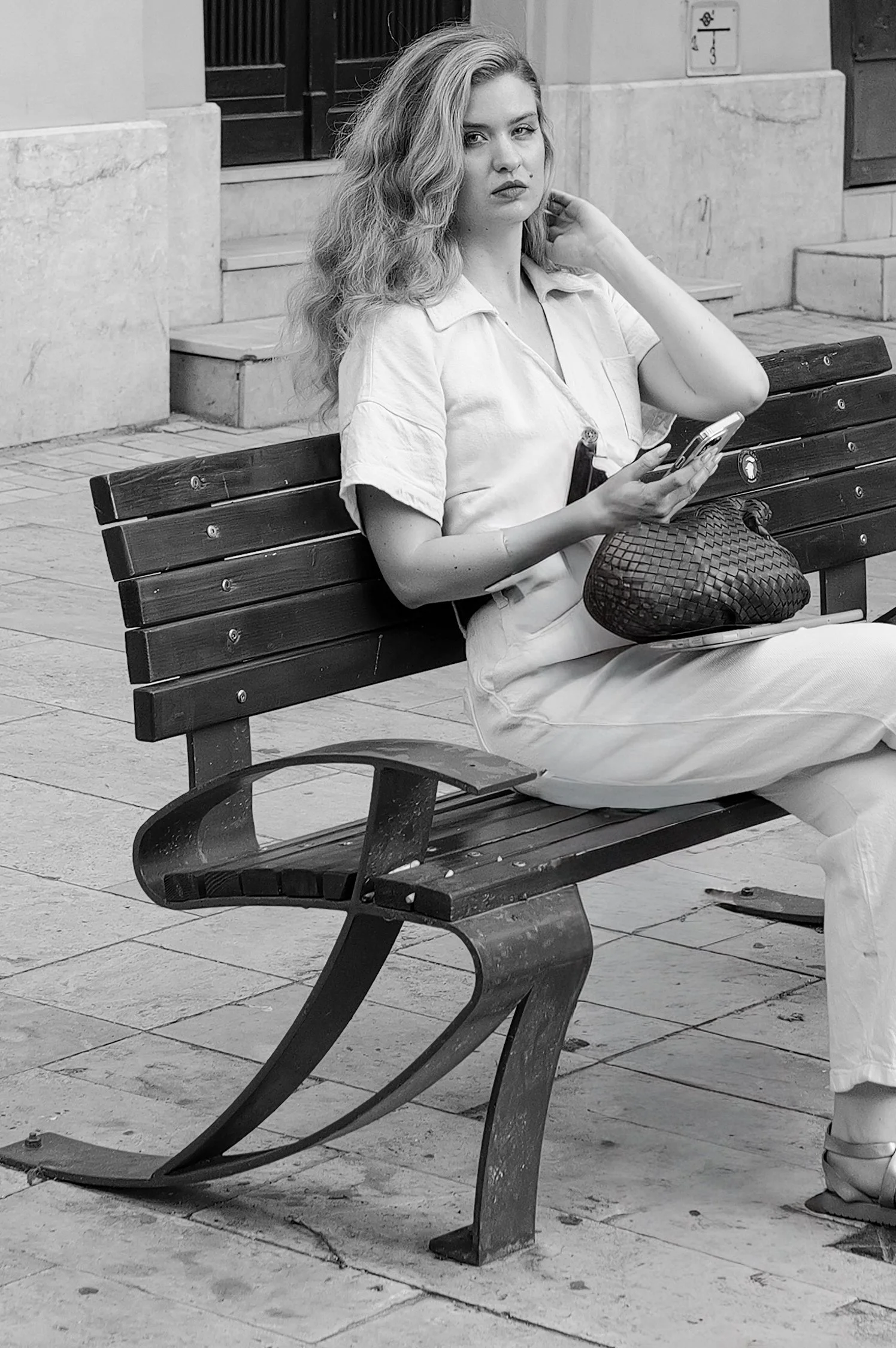 Une femme assise sur un banc en bois noir et métal, tenant un téléphone, avec un sac en bandoulière, dans un environnement urbain.