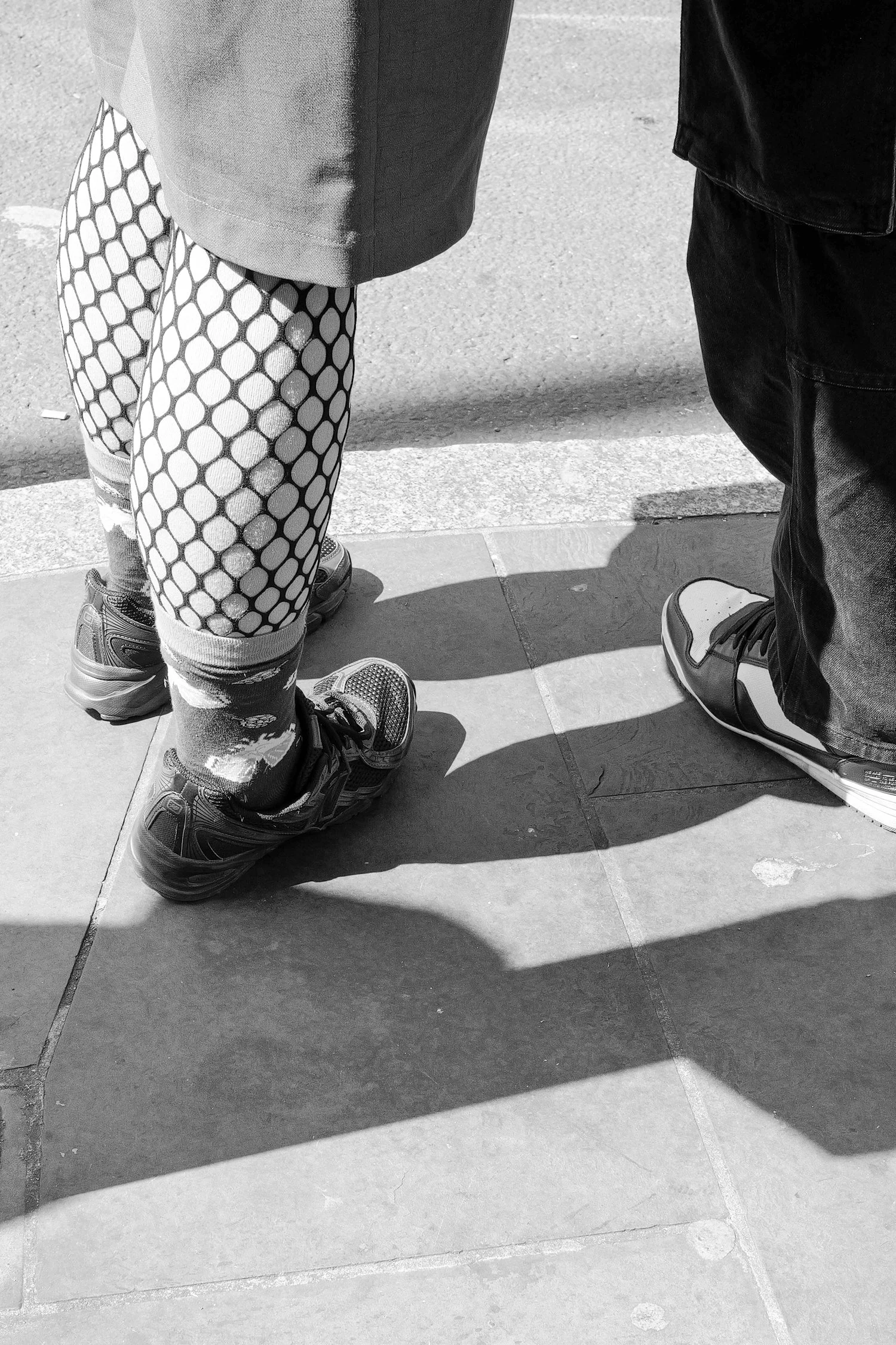 Deux personnes debout sur un trottoir, vue en gros plan de leurs jambes et chaussures, avec ombres projetées sur le sol.