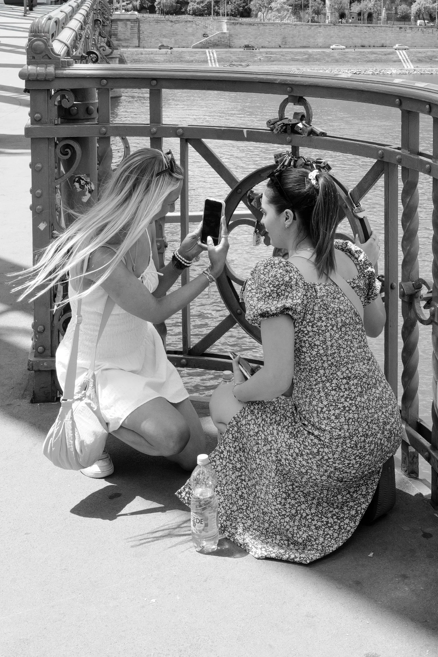 Deux femmes discutent près d'un canal, l'une tenant leur téléphone, avec des cadenas attachés à une barrière en fer, en arrière-plan des voitures et un bâtiment.