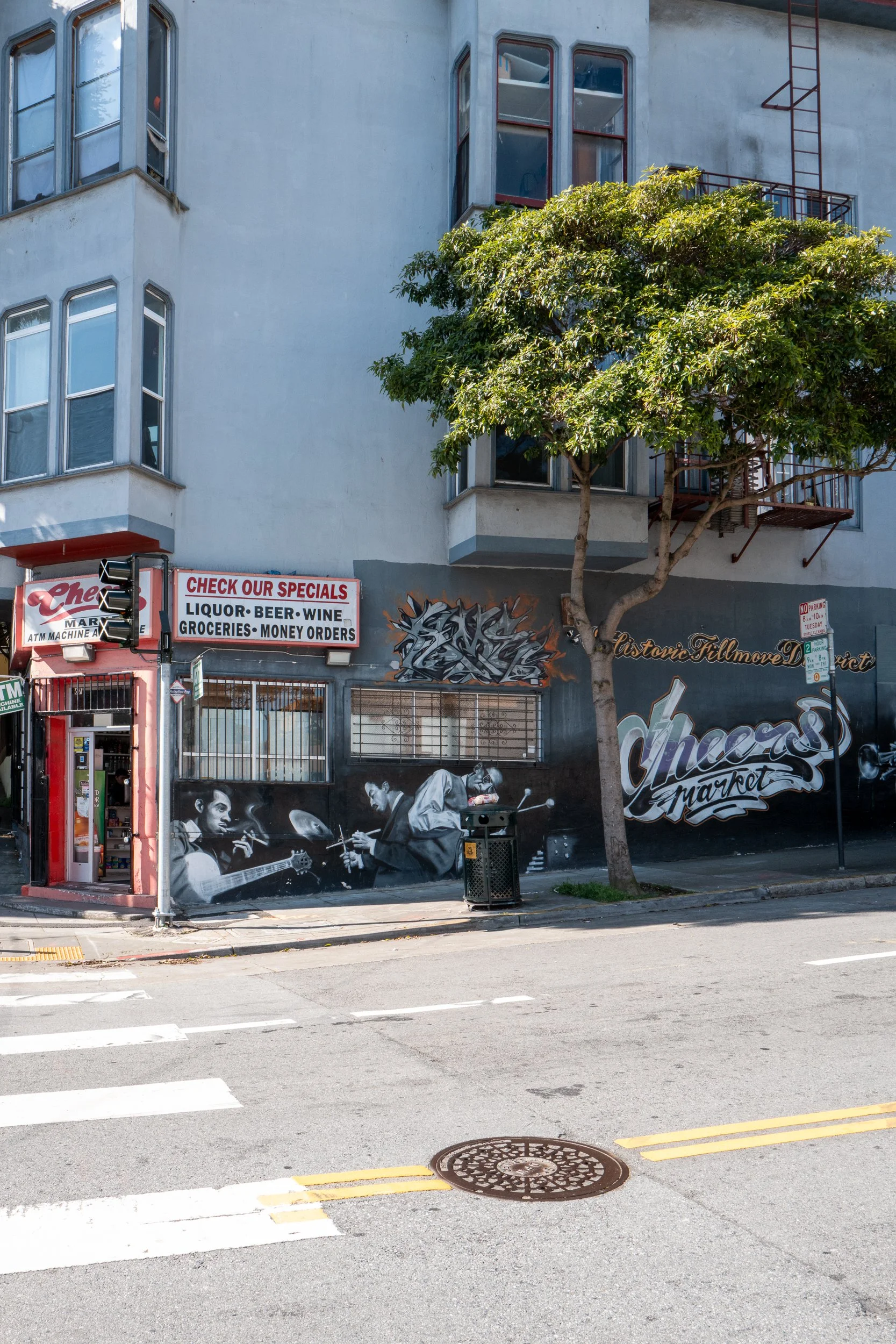 Murale de trois musiciens jouant de la musique à l'extérieur d'une épicerie, avec arbre et bâtiment ancien en arrière-plan, dans une ville