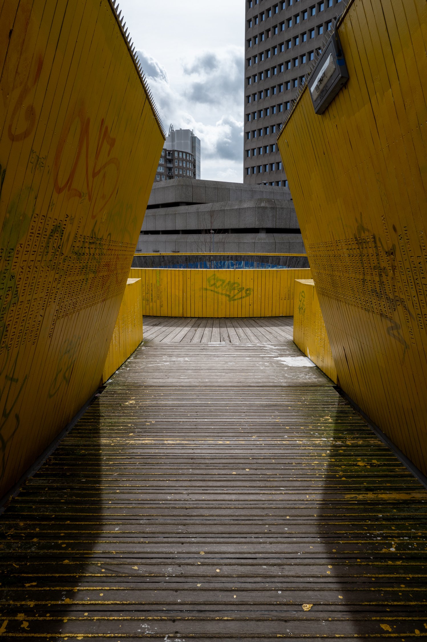 Vue à travers une ouverture formée par deux panneaux en bois jaunes vers un espace public, avec un sol en bois et des bâtiments modernes en arrière-plan, ciel nuageux.