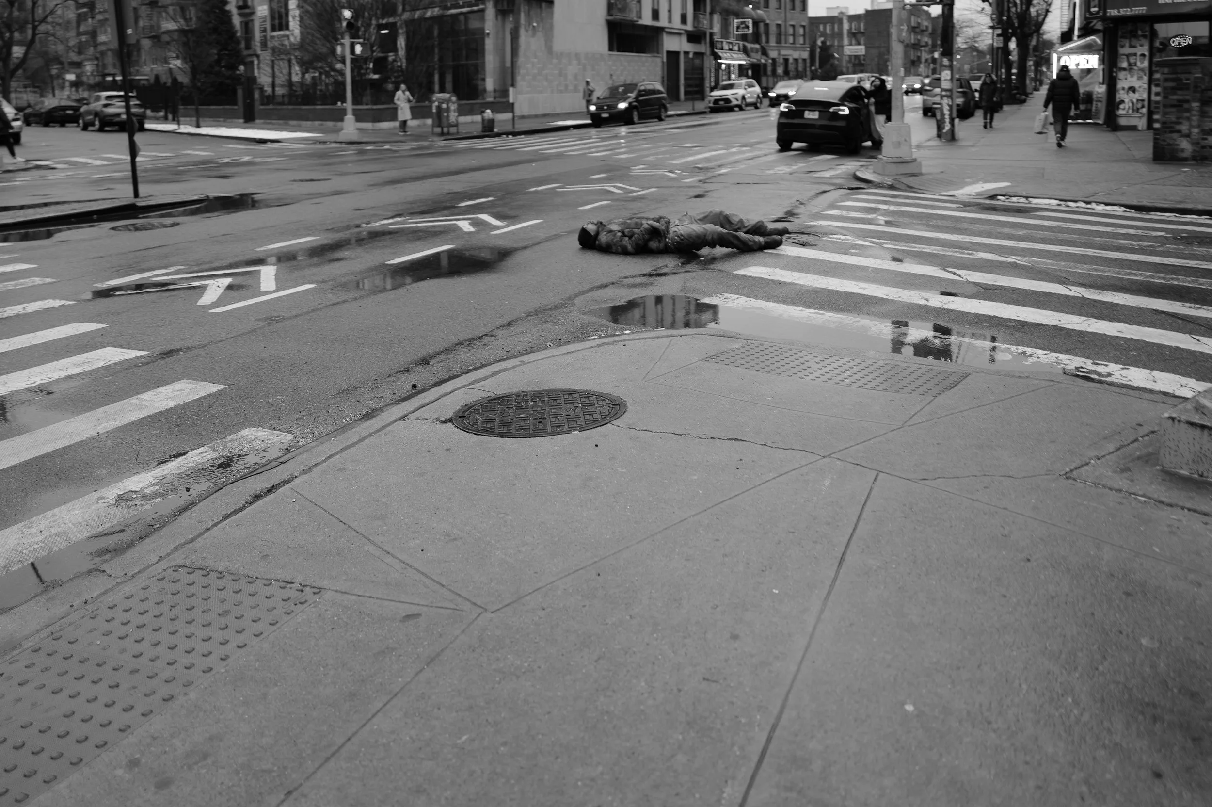 Un homme allongé sur un passage piéton en ville, entouré de voitures et de passants, dans une ambiance sombre et pluvieuse.