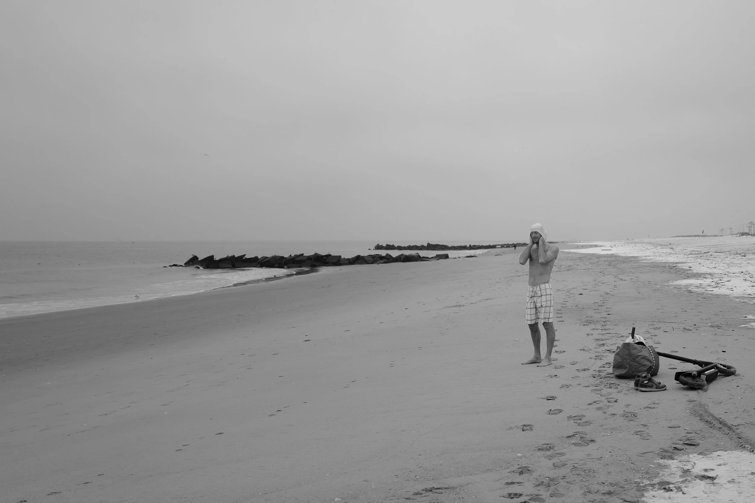 Un homme debout sur une plage, parlant au téléphone, avec des sacs et un scooter électrique à côté, ciel nuageux.