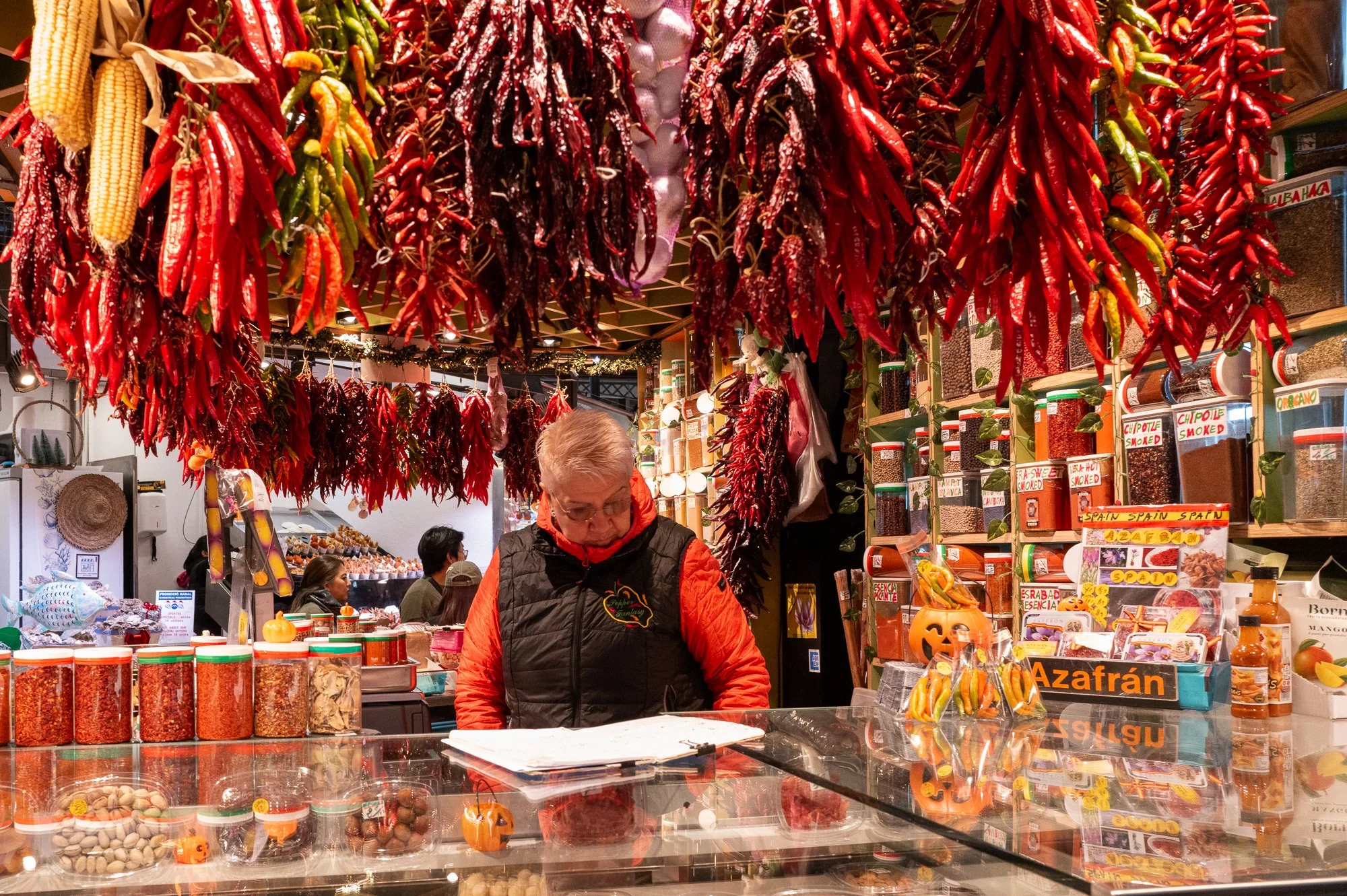 Vendeur devant un stand de marché avec des guirlandes de piments rouges suspendus au plafond, rayonnages de bocaux d'épices et d'assaisonnements, décorations de Halloween, et plusieurs clients en arrière-plan.