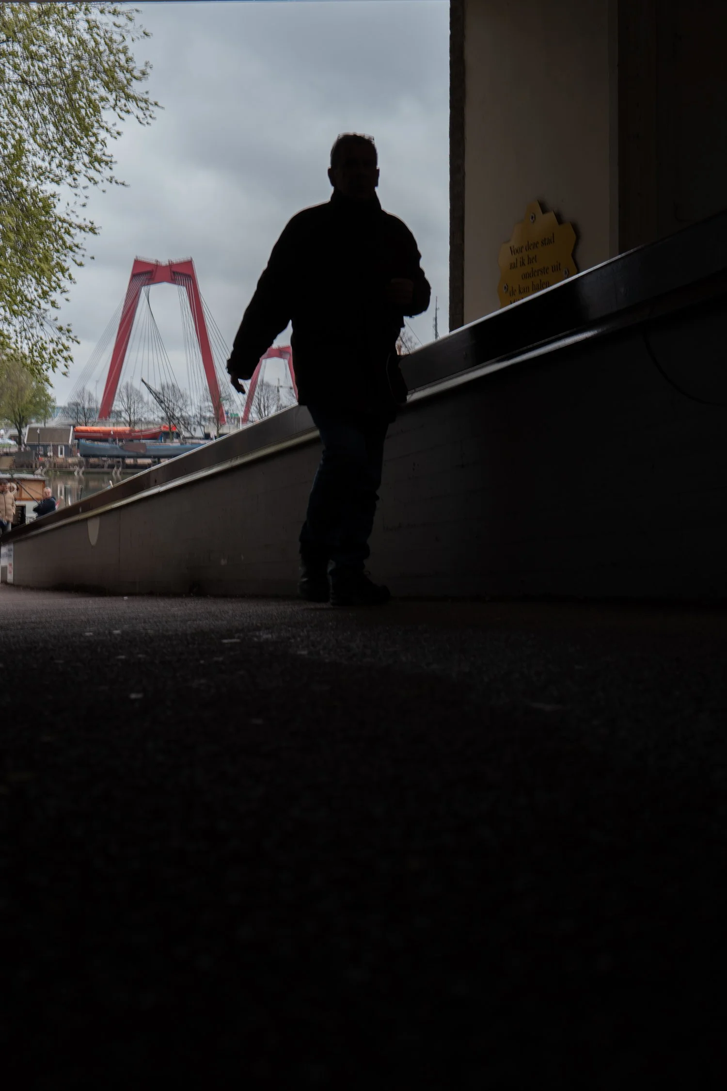 Silhouette d'une personne debout dans un passage sombre, avec le pont rouge en arrière-plan et un ciel nuageux.
