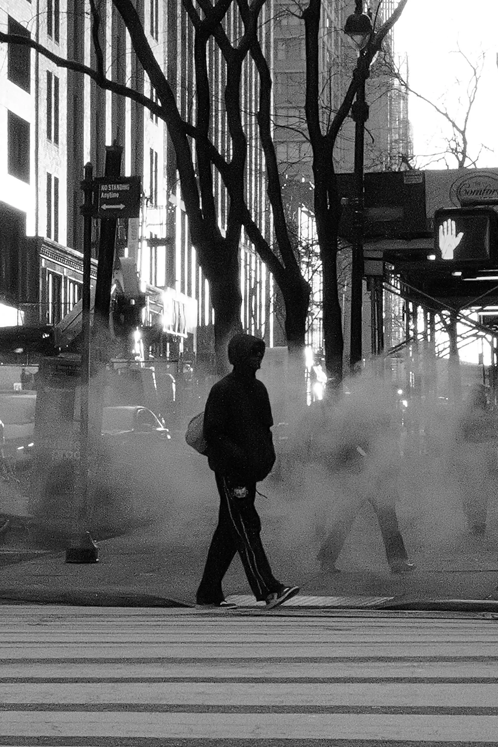 Une silhouette d'une personne marchant à un passage pour piétons dans une rue urbaine en noir et blanc. Il y a de la fumée ou de la vapeur près du sol et des arbres sans feuilles sur le trottoir.