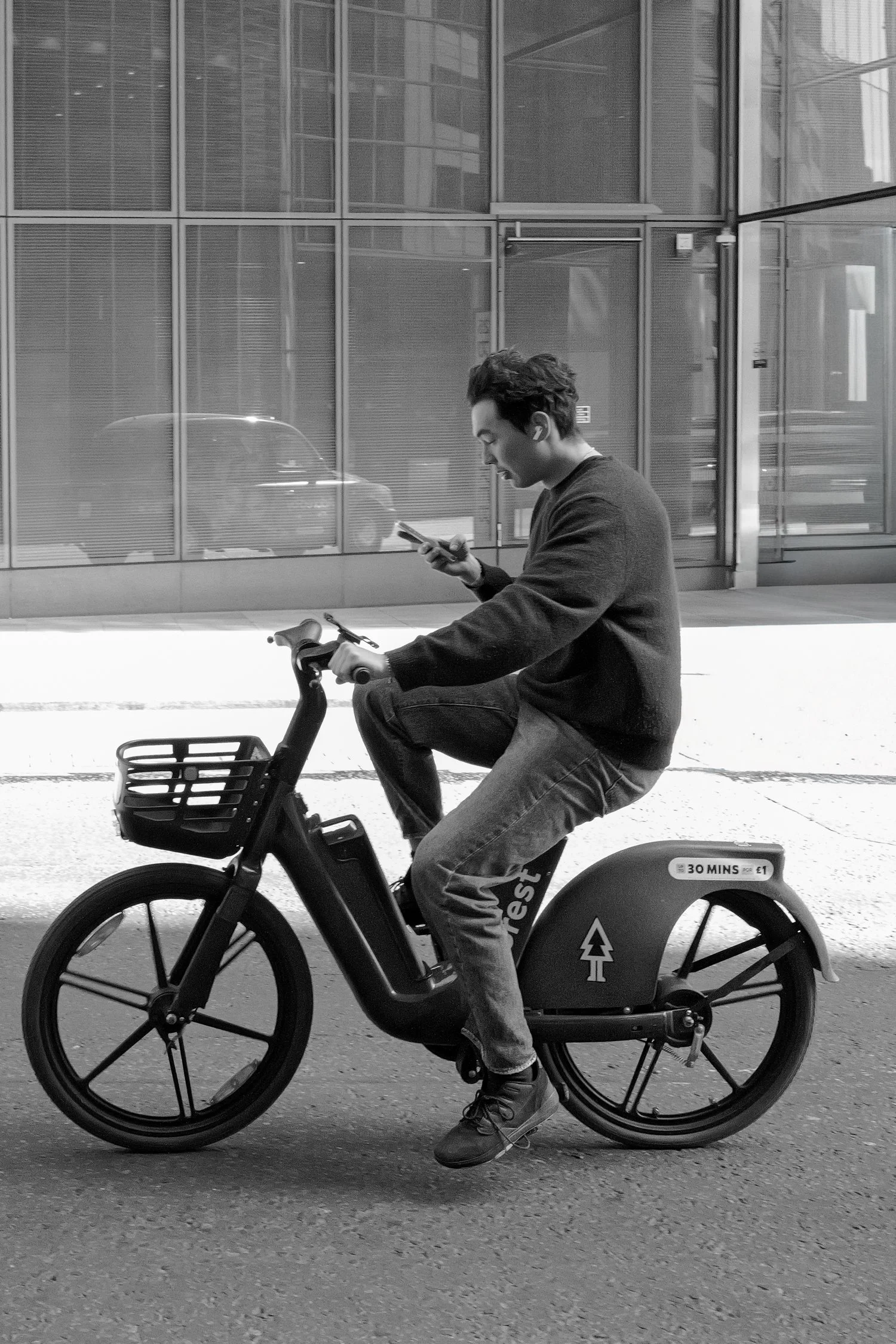 Un homme à vélo électrique en ville, regardant son téléphone, vêtement noir, dans un environnement urbain moderne avec de grands bâtiments en arrière-plan.