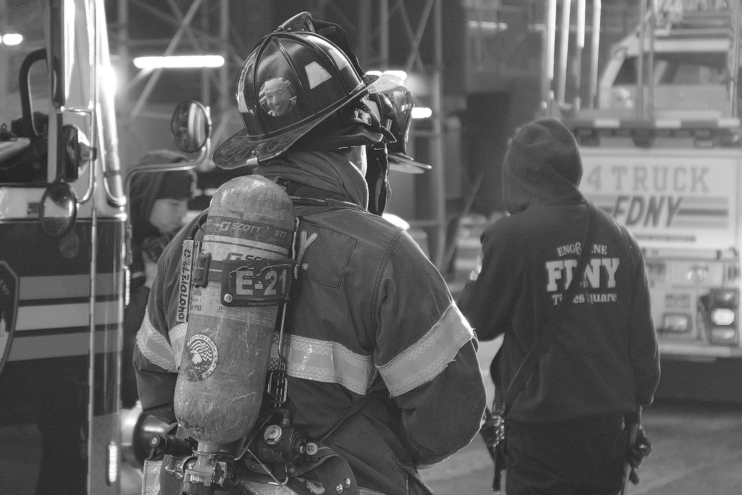 Un pompier en uniforme avec un casque et une bouteille d'oxygène sur le dos, en discussion avec un autre individu portant une veste avec l'inscription 'FIRE DEPT' sur le dos, dans un environnement industriel ou d'urgence.