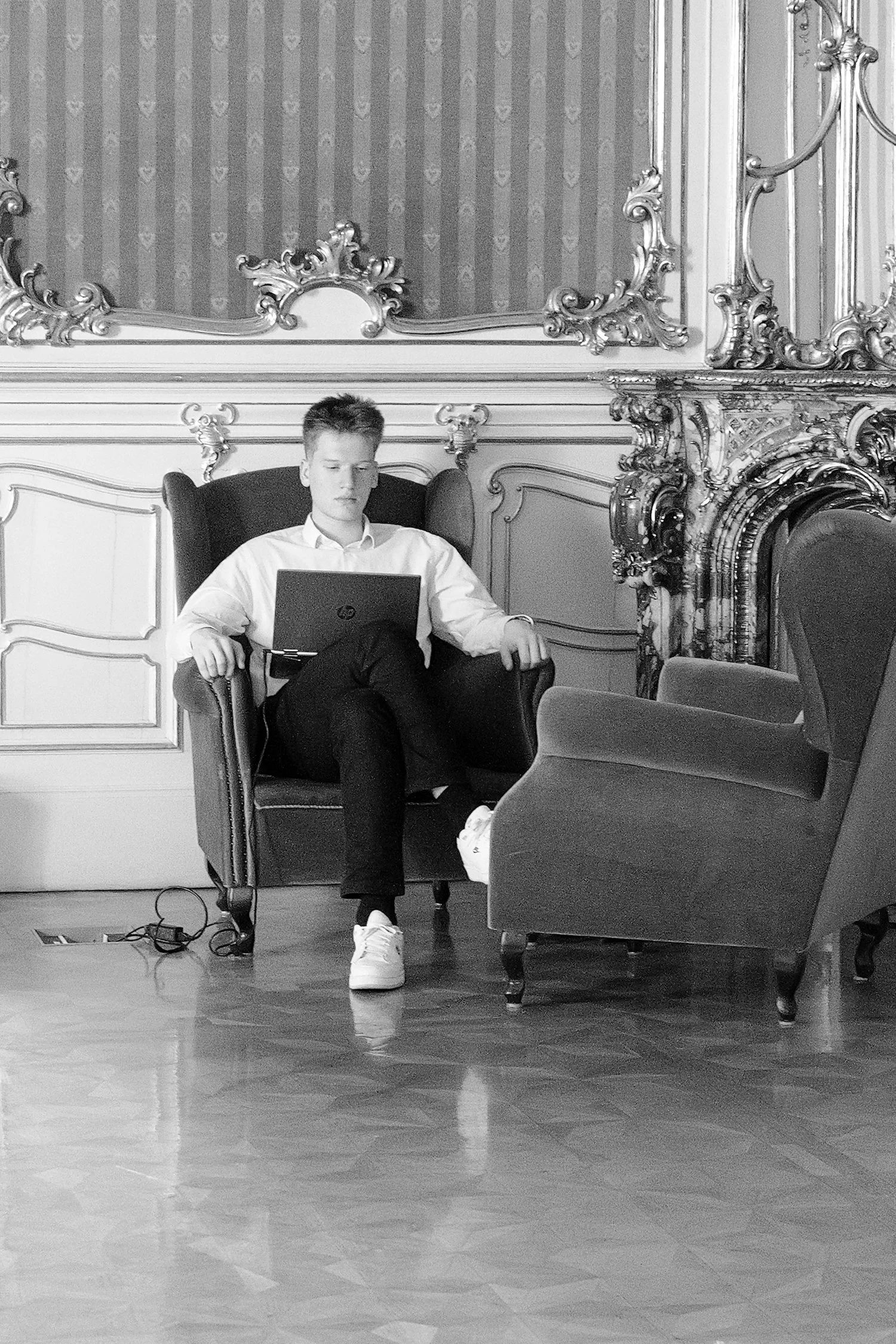 Un jeune homme assis dans un fauteuil en bois, portant un pantalon noir, une chemise blanche, des baskets blanches, dans une pièce ornée de décoration classique et un mur avec un motif à rayures. Il regarde son ordinateur portable sur ses genoux et s