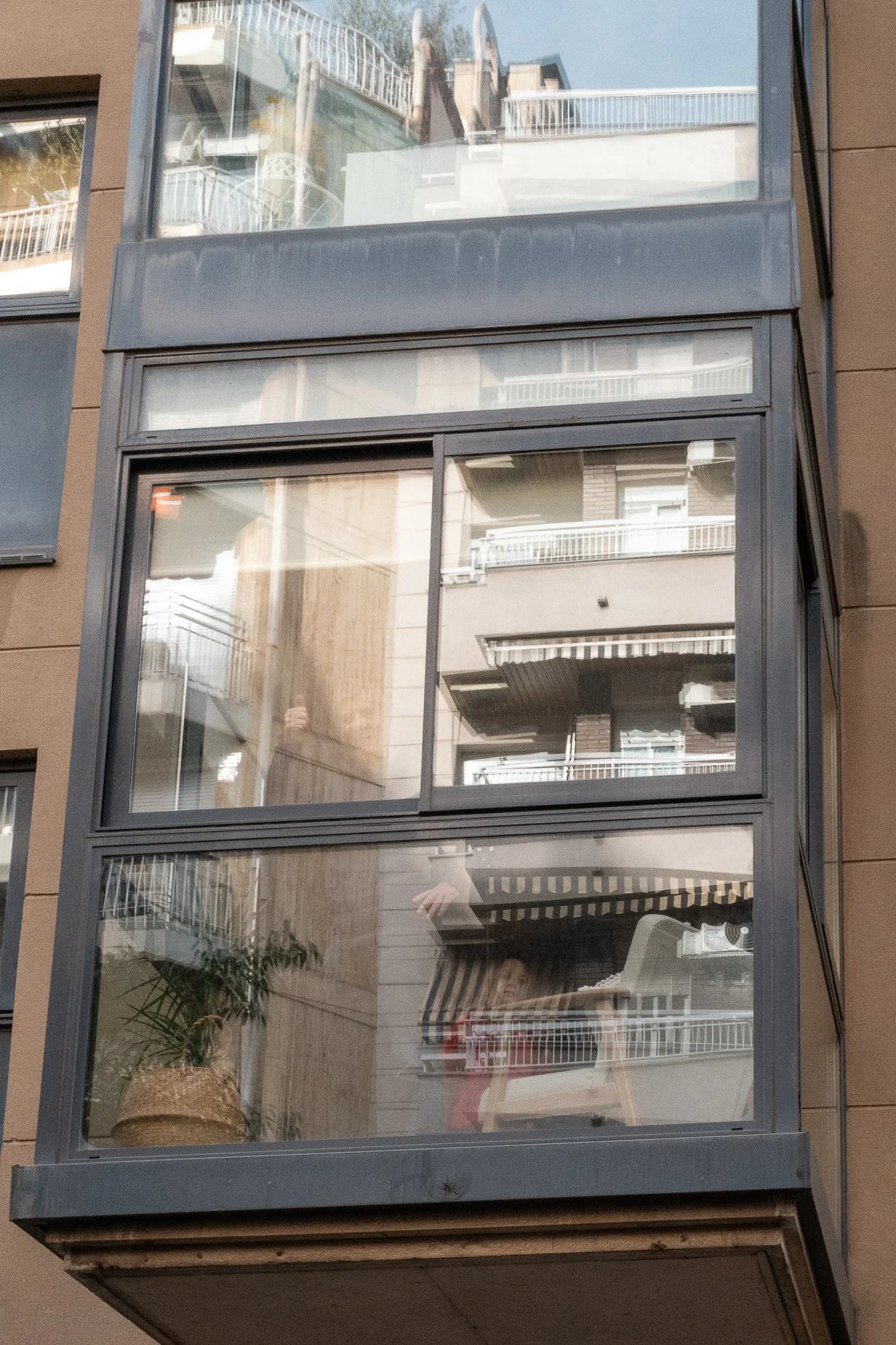 Vue d'un balcon en verre avec une plante en pot et un tapis à rayures, reflet d'autres balcons d'immeubles.