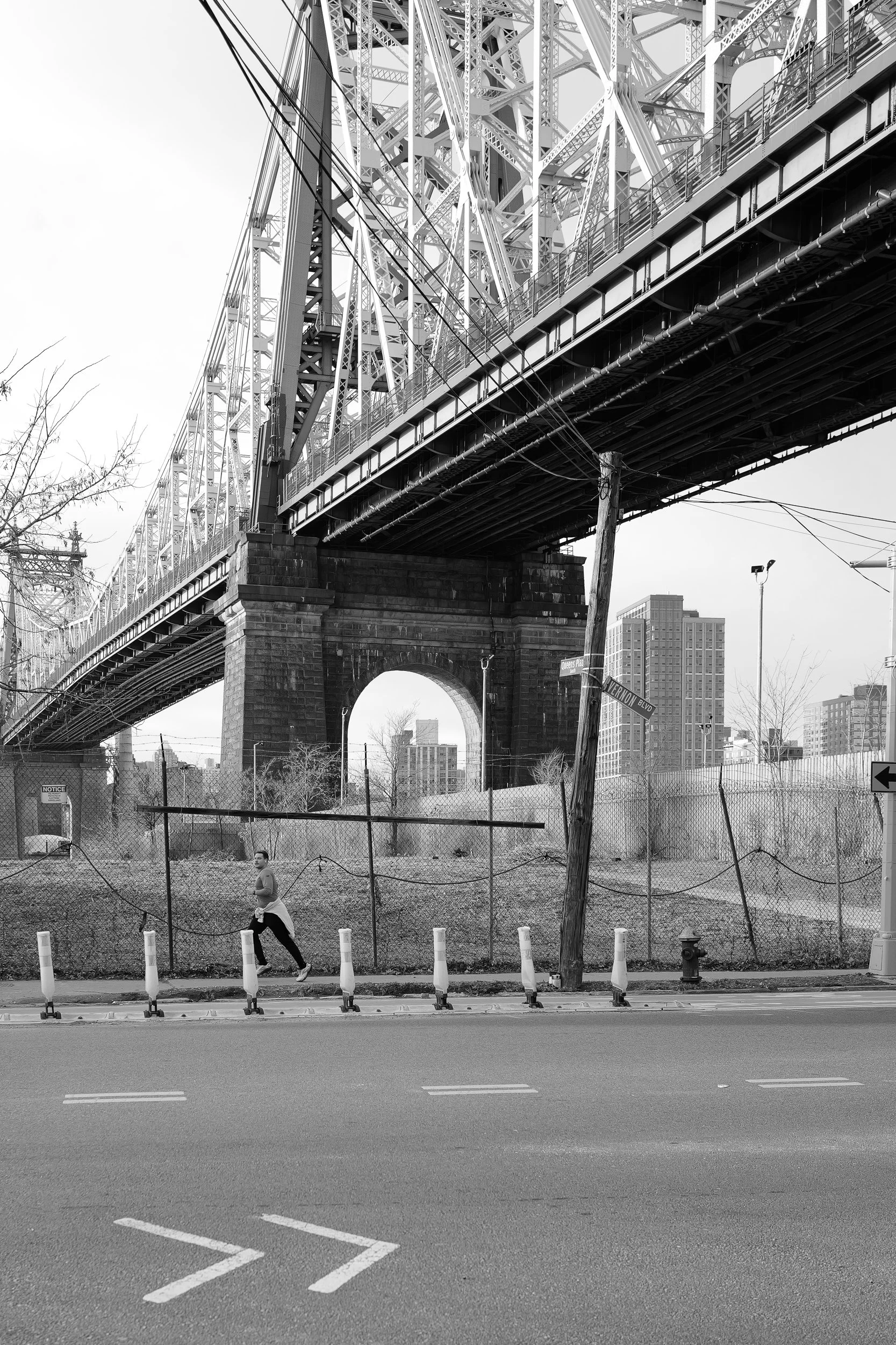 Photo en noir et blanc d'un pont suspendu avec structure en métal, vue de dessous, avec un homme marchant à côté d'une clôture et un poteau électrique penché, ville en arrière-plan.