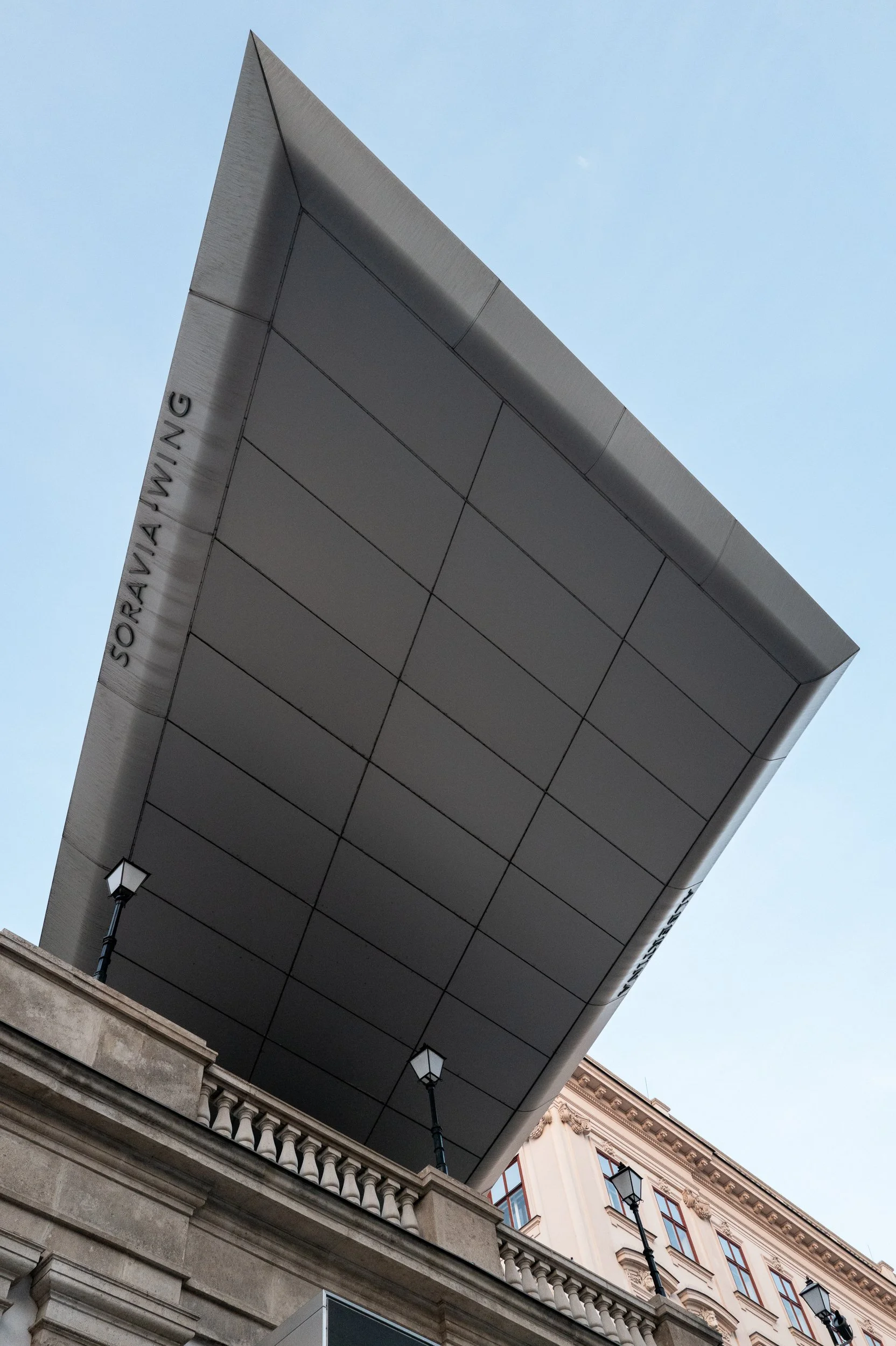 Vue en contre-plongée d'un bâtiment moderne avec une grande structure en surplomb argentée et des lampadaires classiques en bas, situé au-dessus d'une architecture ancienne.