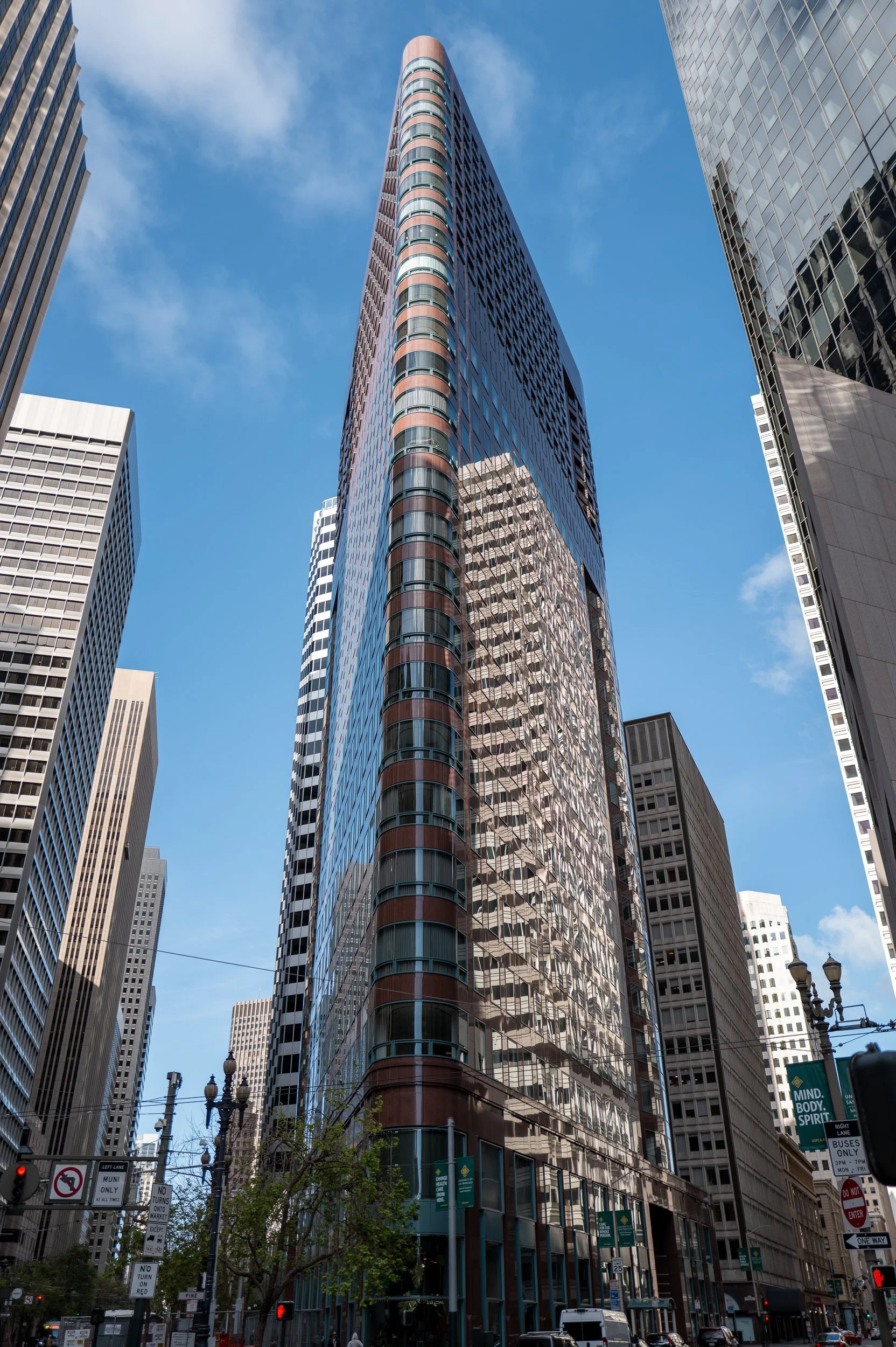 Une grande tour en verre dans une ville, entourée d'autres gratte-ciel, avec un ciel bleu clair et quelques nuages.