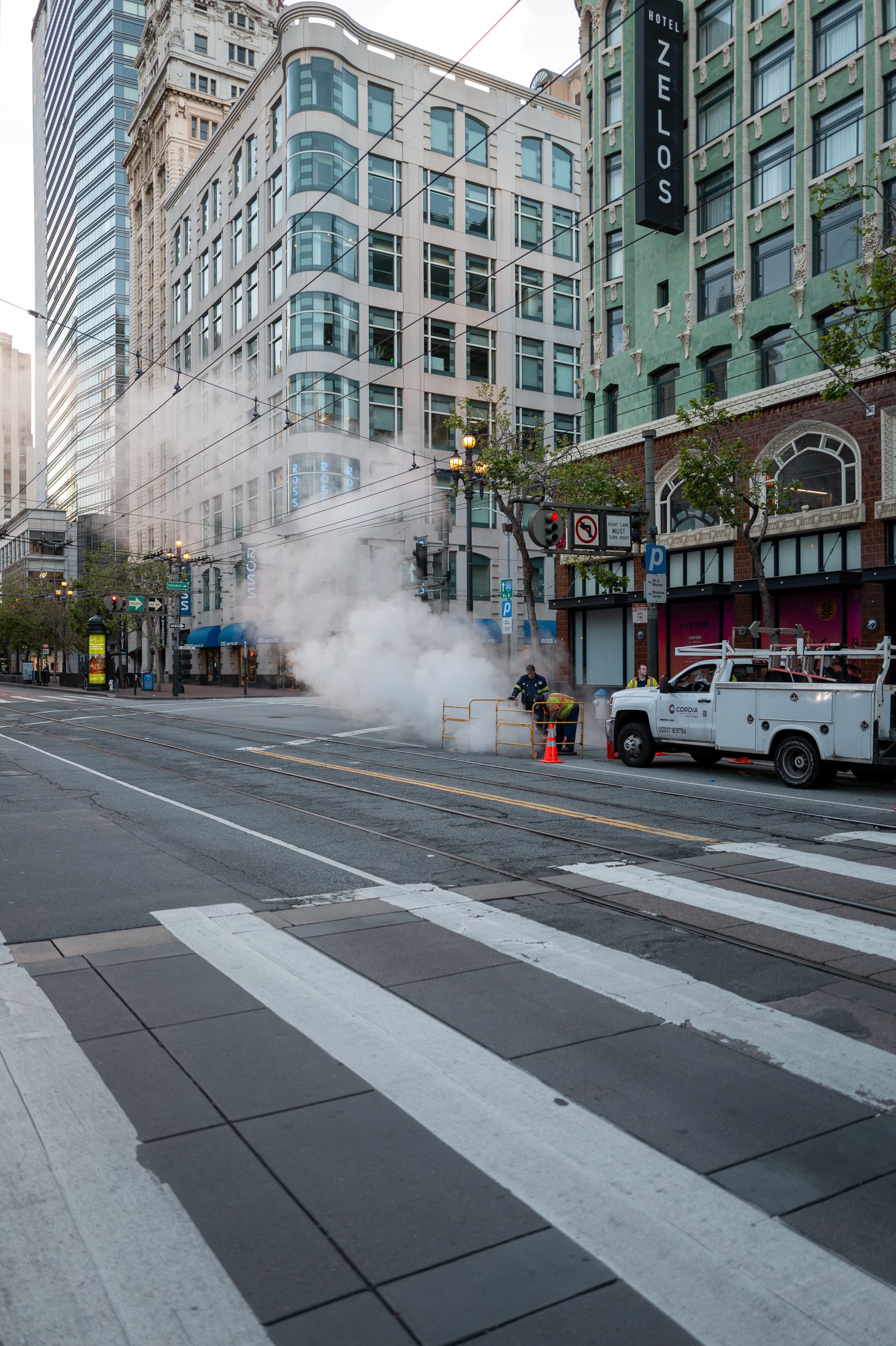 Travaux avec vapeur sortant du sol sur une rue urbaine, avec des bâtiments modernes et un véhicule de maintenance