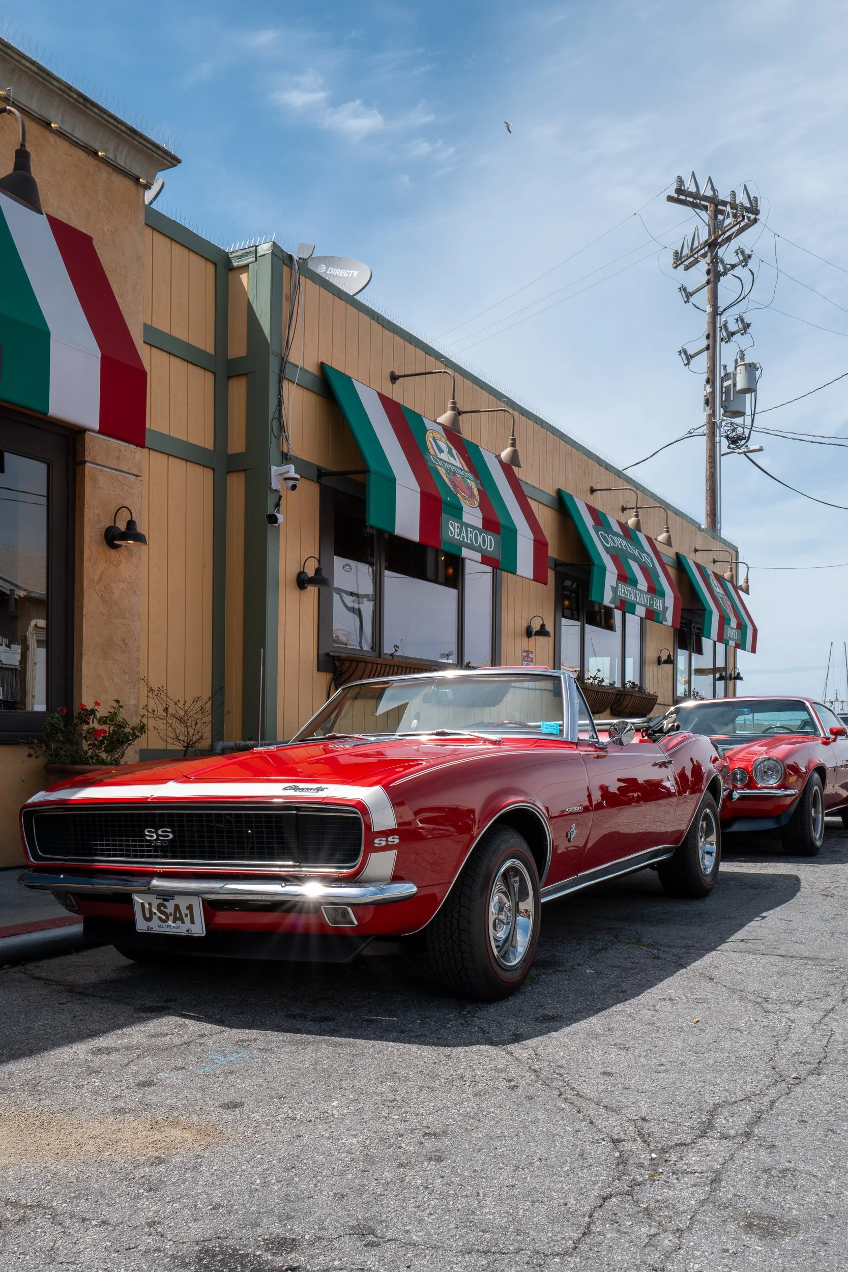 Deux voitures classiques rouges stationnées devant un restaurant en bord de mer avec des enseignes en rayures vertes, blanches et rouges, sous un ciel bleu avec quelques nuages et un oiseau en vol.