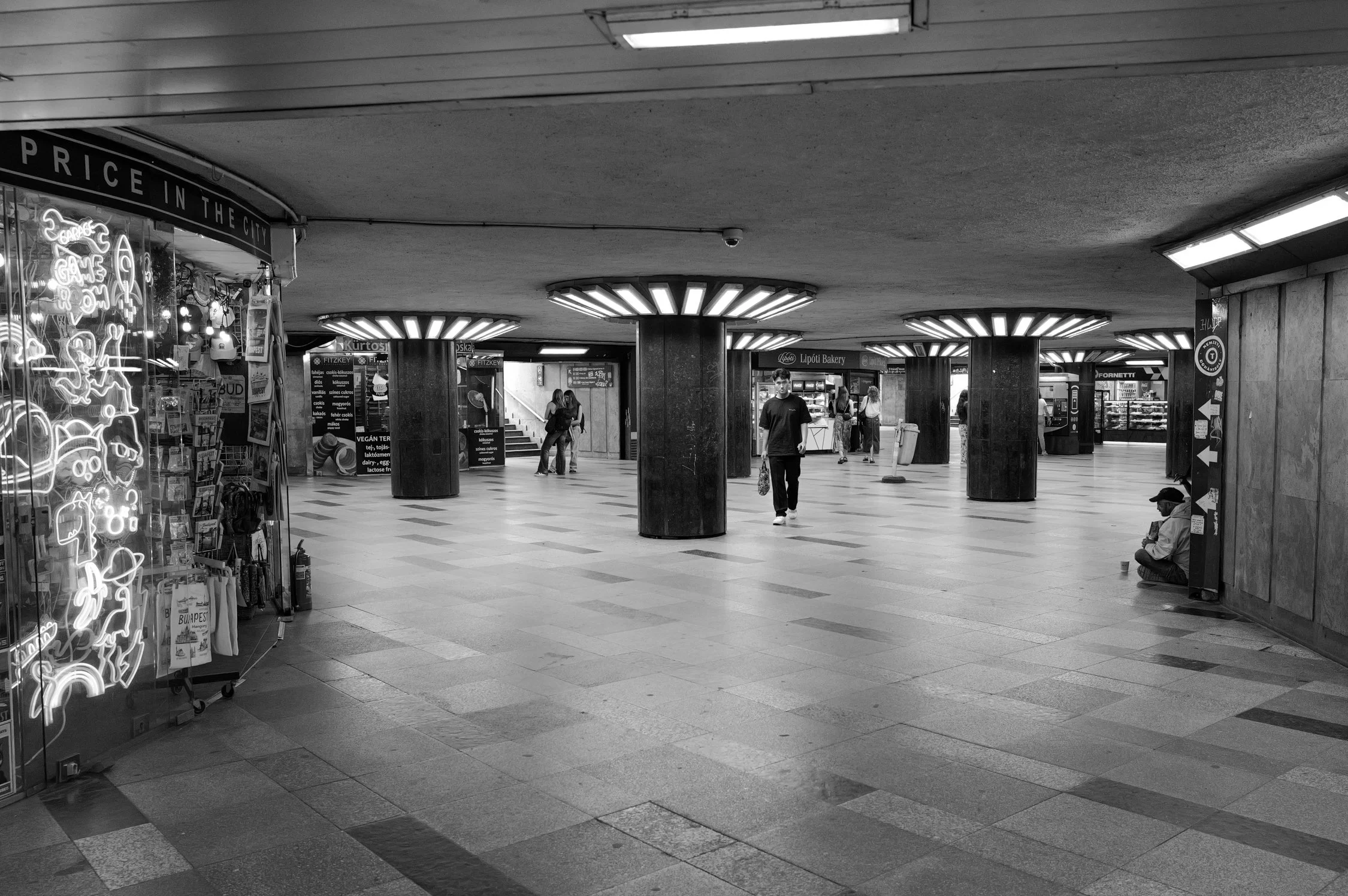 Une photo en noir et blanc d'une station de métro souterraine avec plusieurs colonnes cylindriques, des boutiques et des passants, dont certains sont assis ou marchent.