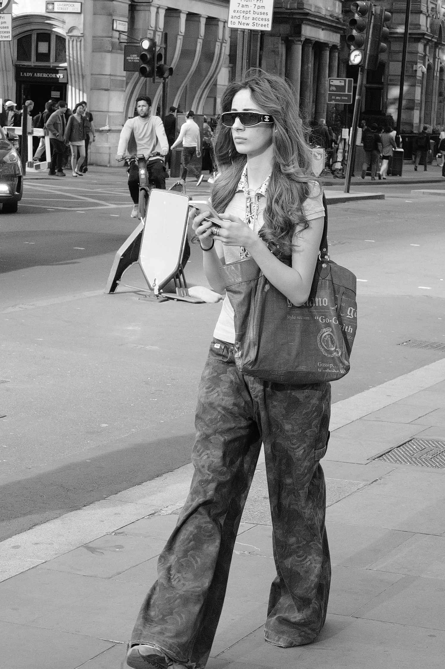 Une femme portant des lunettes de soleil, un t-shirt léger et un sac à bandoulière marche dans une rue urbaine, concentrée sur son téléphone. En arrière-plan, des passants, des voitures et des bâtiments historiques sont visibles.