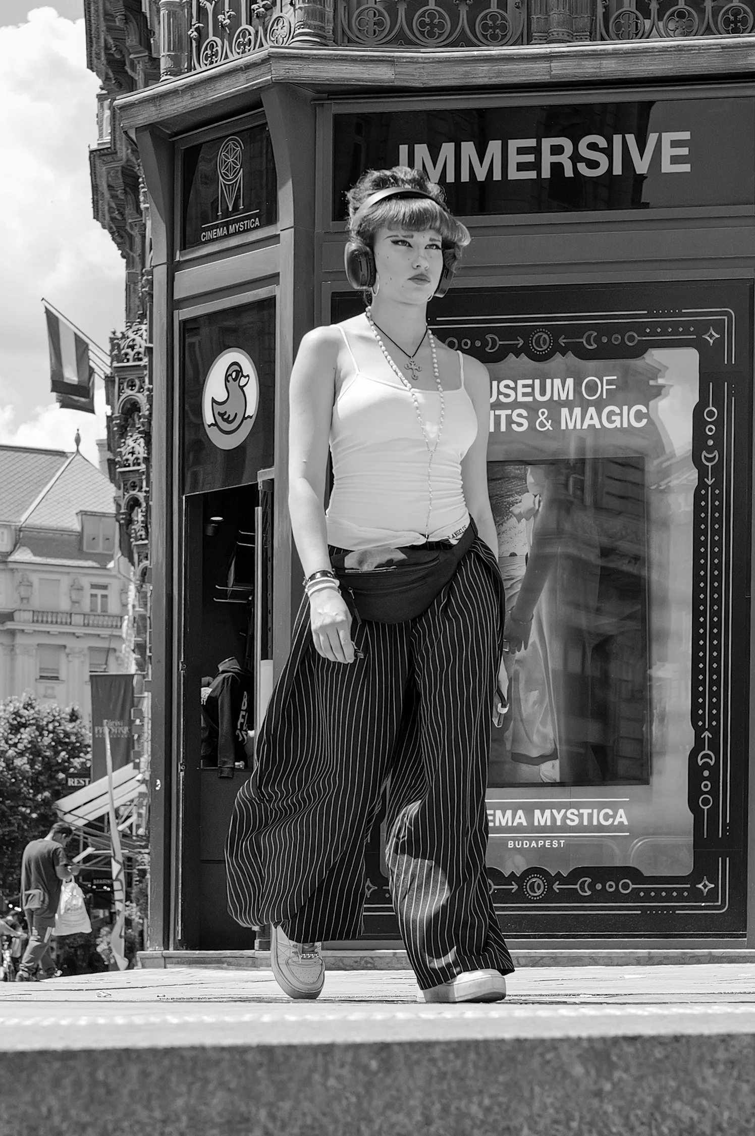 Une femme marche dans une rue urbaine portant des écouteurs, une chemise blanche, un collier, un bracelet et un sac à la main, devant un panneau d'affichage avec des textes en anglais, dans un environnement citadin avec des bâtiments visibles en arri