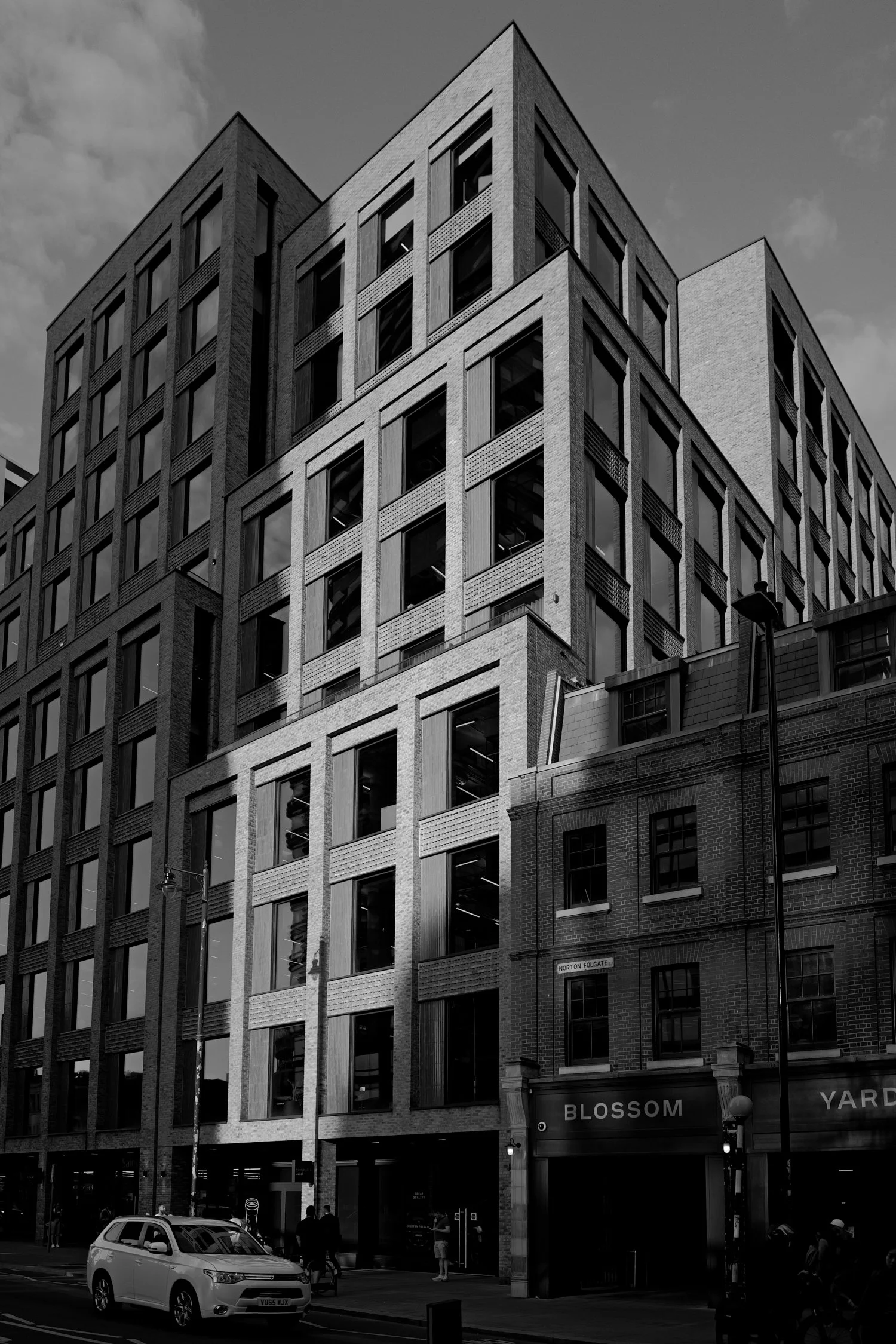 Un immeuble moderne en pierre et en verre avec des fenêtres rectangulaires, situé en ville. Il y a une voiture blanche stationnée au premier plan et des personnes marchant sur le trottoir. Signalisation indiquant 'Norton Folgate' et 'Blossom Yard'.