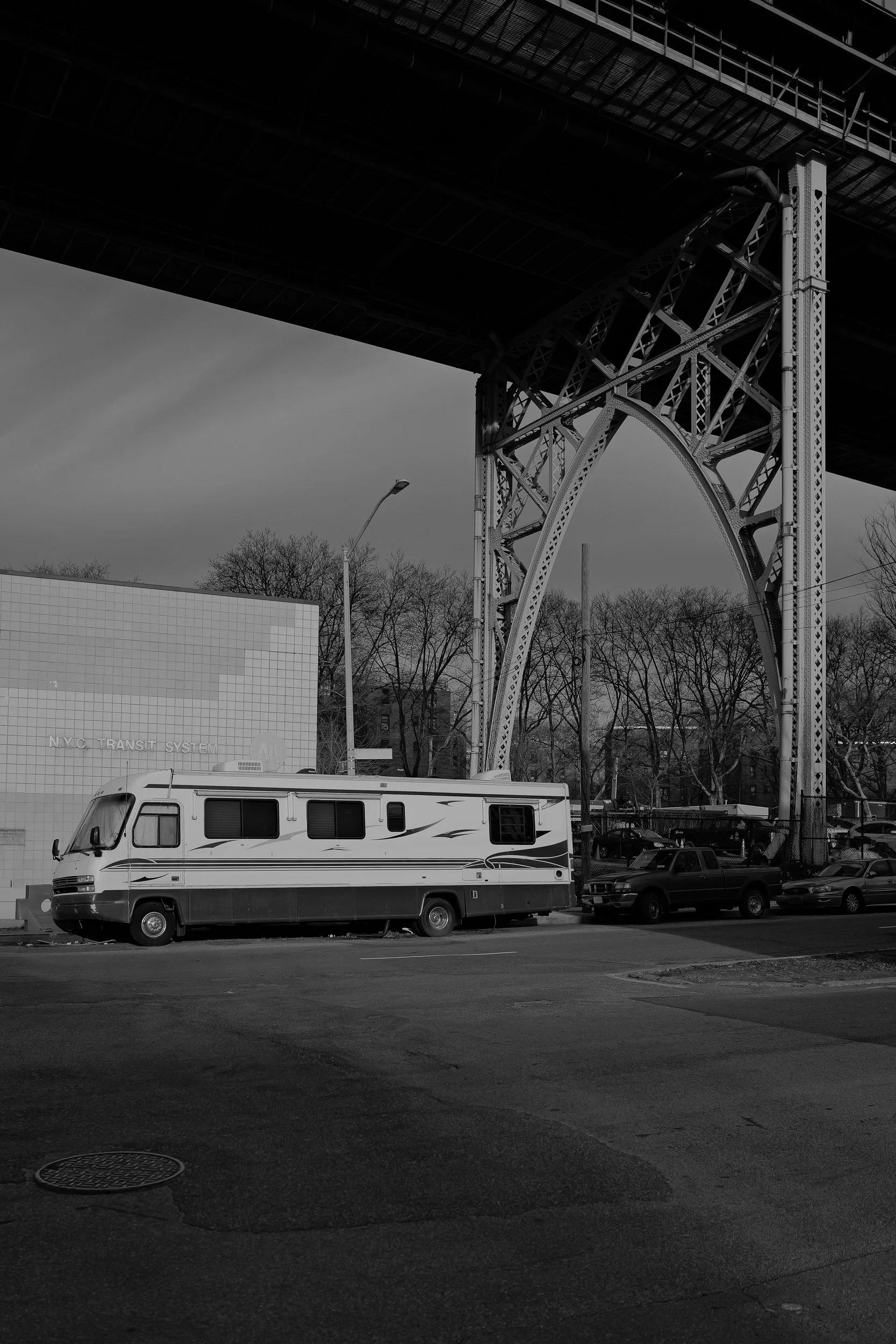 Un camping-car stationné sous un pont, avec des voitures à côté, un mur avec un motif de carré et des arbres en arrière-plan. La scène est en noir et blanc.