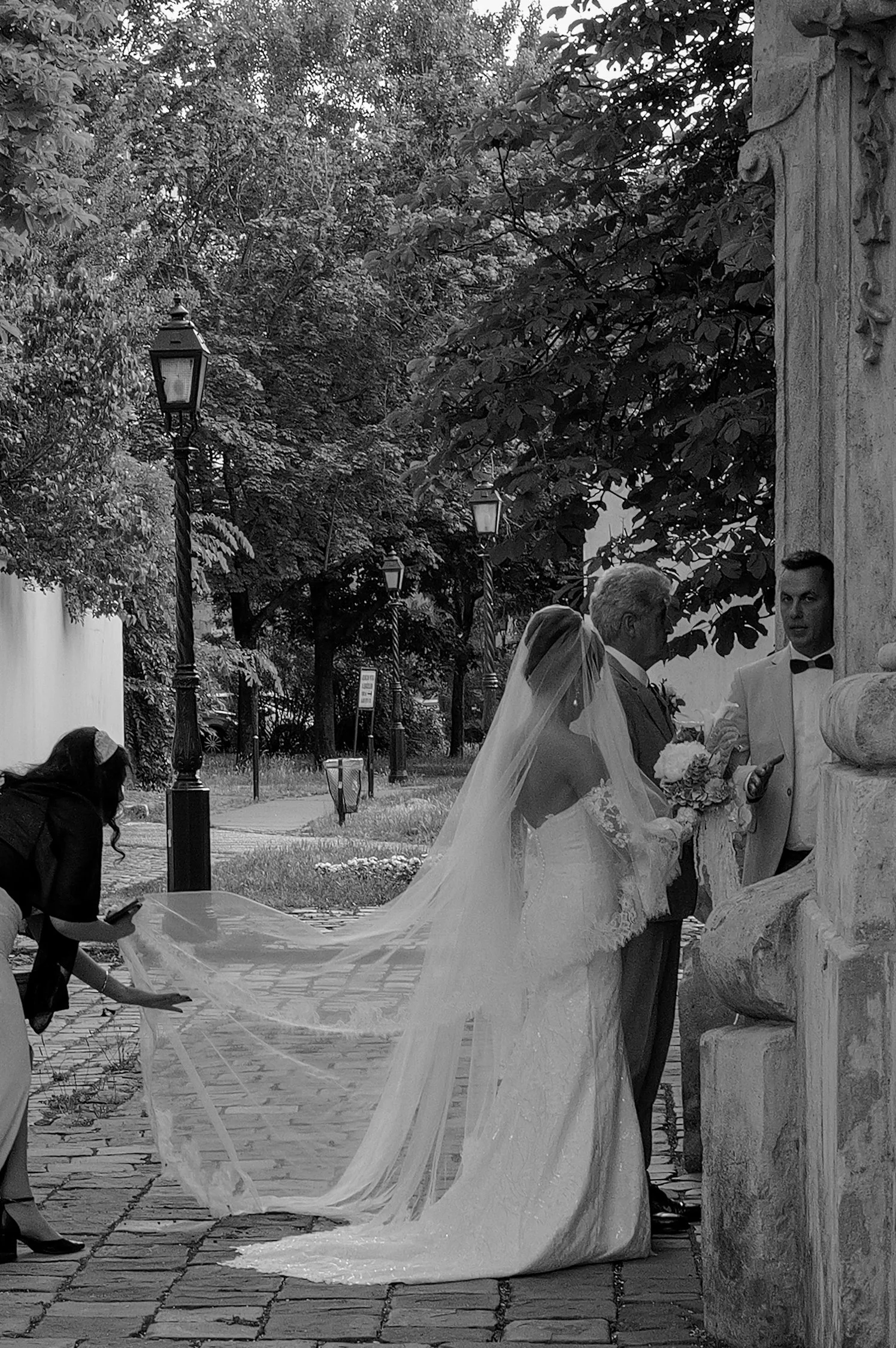 Une scène de mariage en noir et blanc où un couple, probablement les mariés, se tient près d'une sculpture ou d'un monument, avec un officiant ou un membre du personnel en uniforme. Une femme portant une robe de mariée avec une longue traîne et un vo