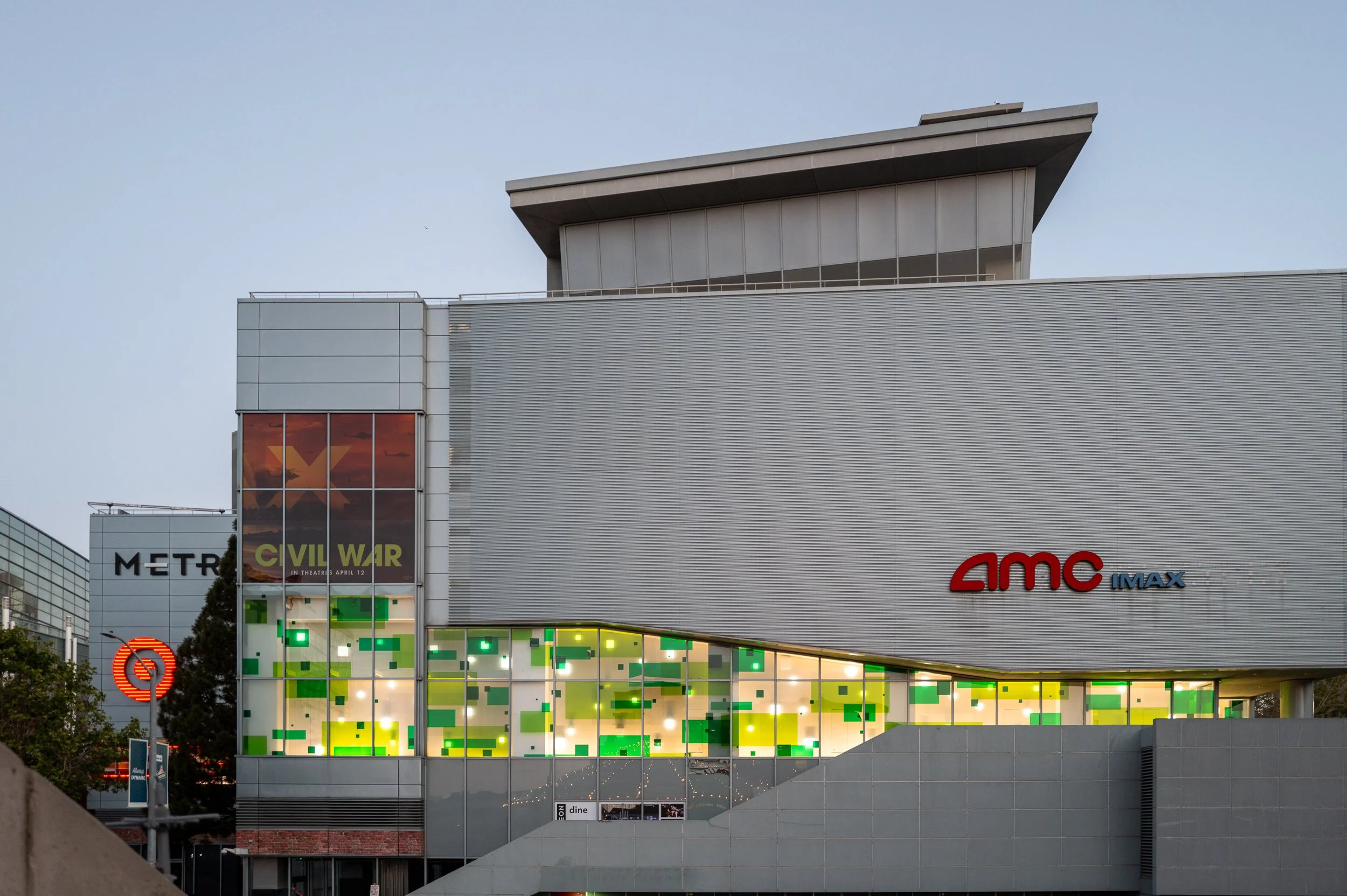 Façade moderne d'une salle de cinéma AMC IMAX avec signage au crépuscule, vitres colorées vert, jaune et orange, et affiches de films, notamment 'Civil War' et un autre film non identifié.