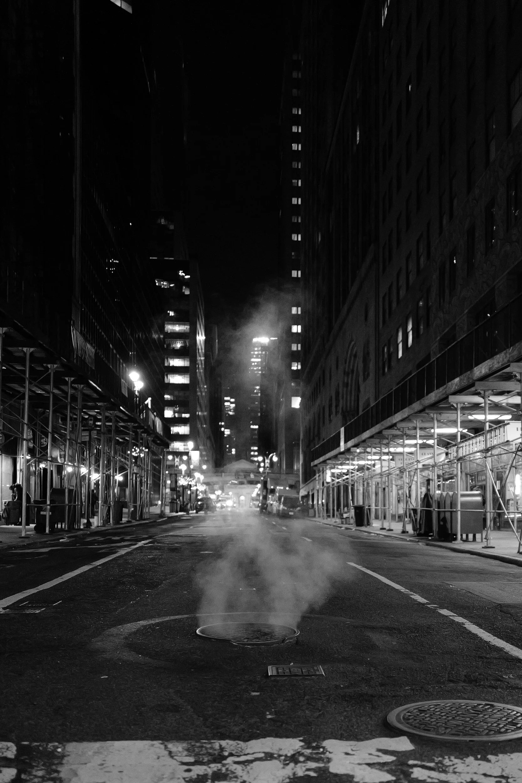 Une rue urbaine vide la nuit, avec des bâtiments hautes, quelques lampadaires, de la fumée s'échappant d'une bouche d'égout, et des échafaudages sur les côtés, en noir et blanc.