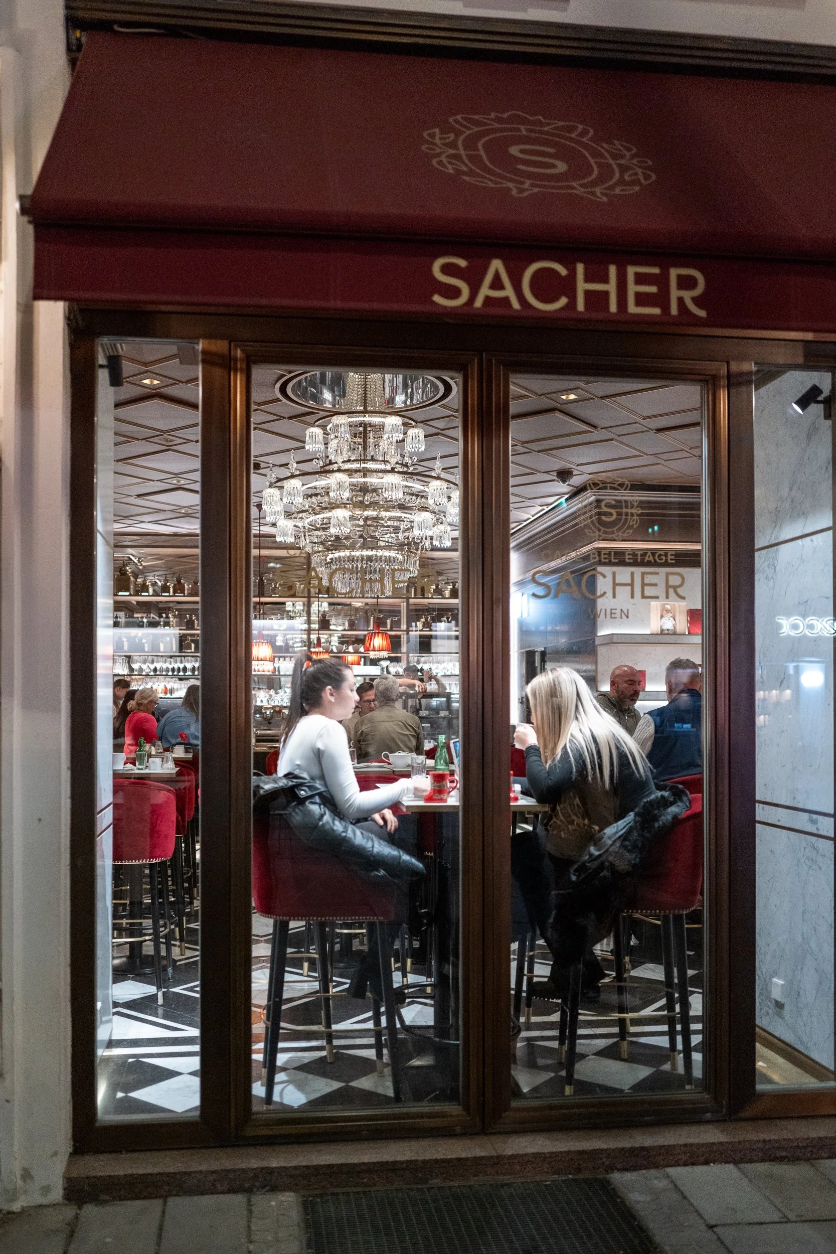 Vue de l'intérieur d'un restaurant nommé Sacher, avec des clients assis au bar, un chandelier au plafond et une décoration élégante.
