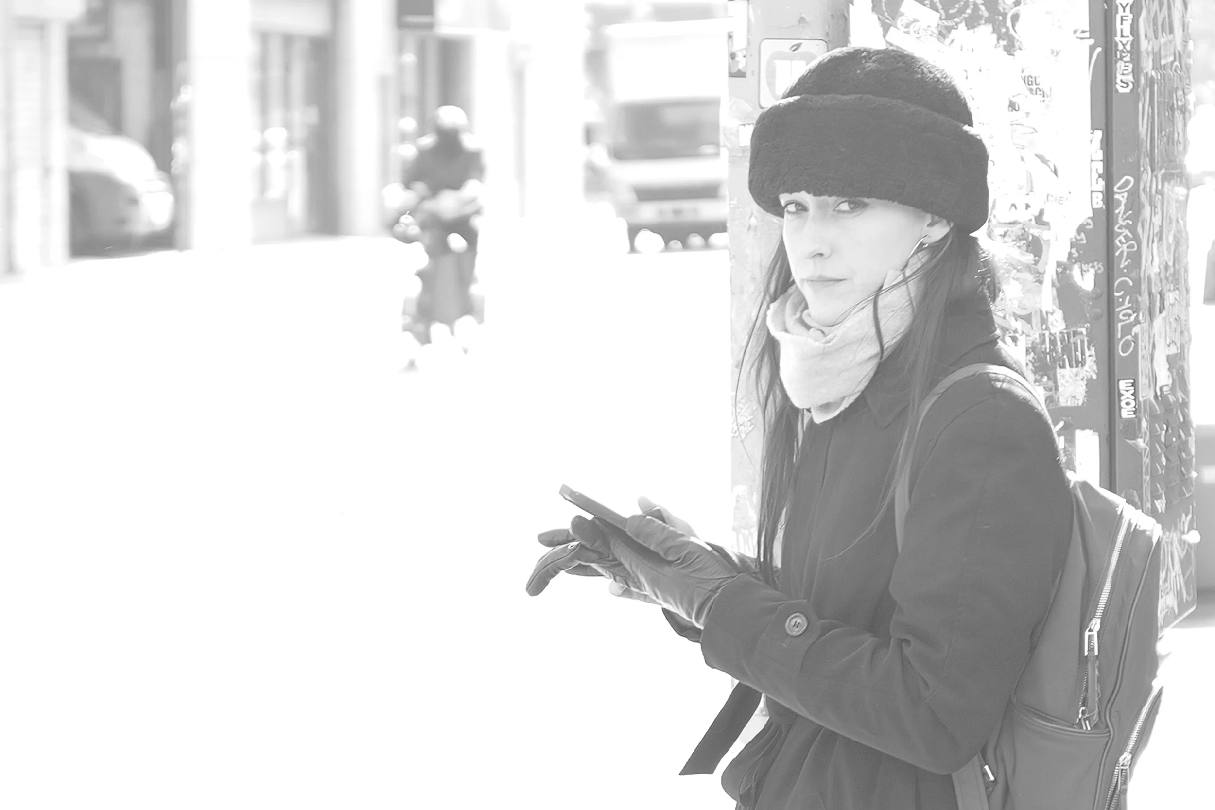Une femme portant un bonnet en peluche noir, un manteau sombre, une écharpe beige, des gants en cuir et un sac à dos regarde son téléphone en dehors, dans une rue ensoleillée.