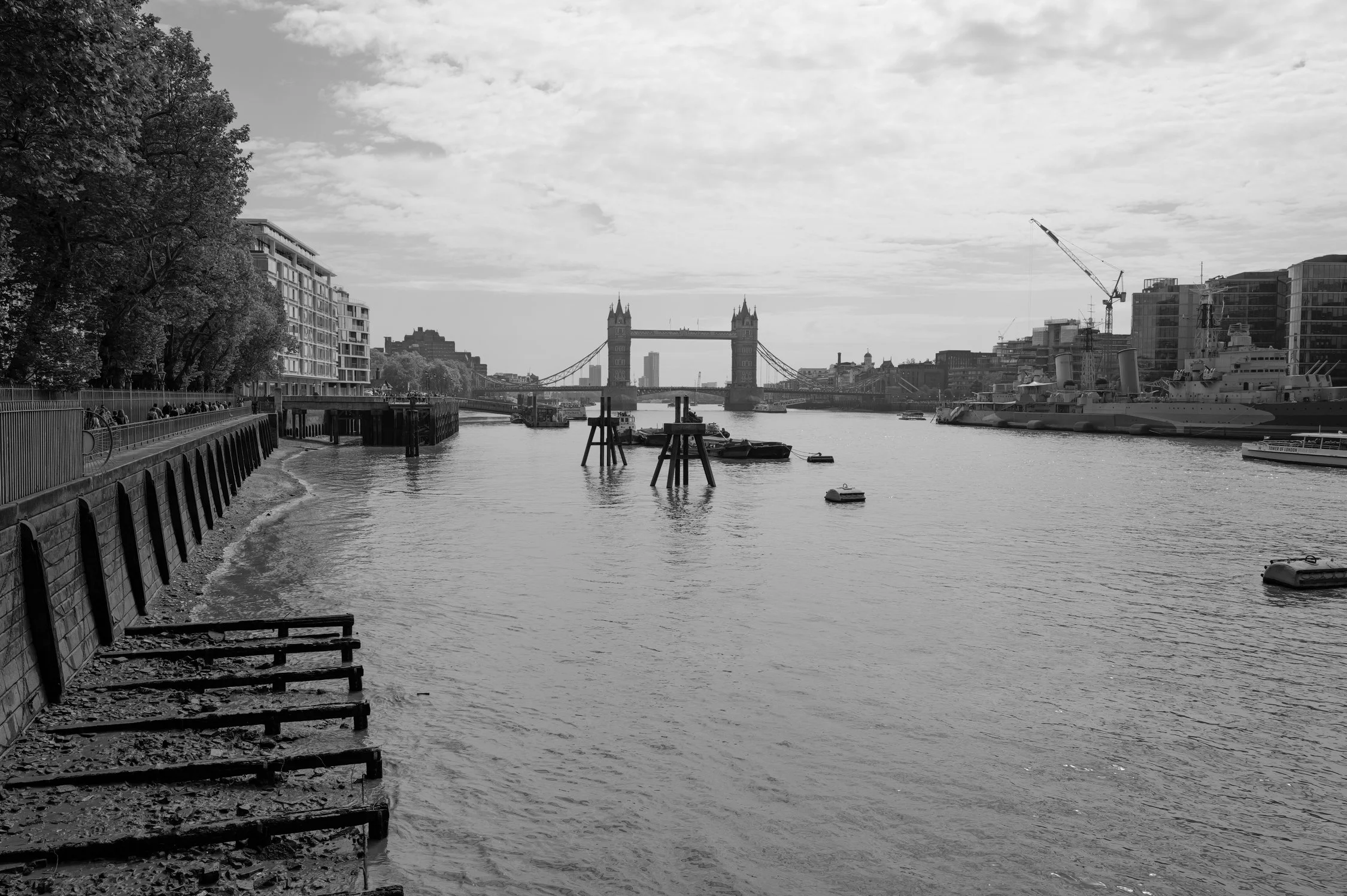 Vue en noir et blanc de la Tamise à Londres avec le pont de Tower Bridge en arrière-plan, des bâtiments modernes bordant la rivière, des bateaux amarrés et un ciel partiellement nuageux.