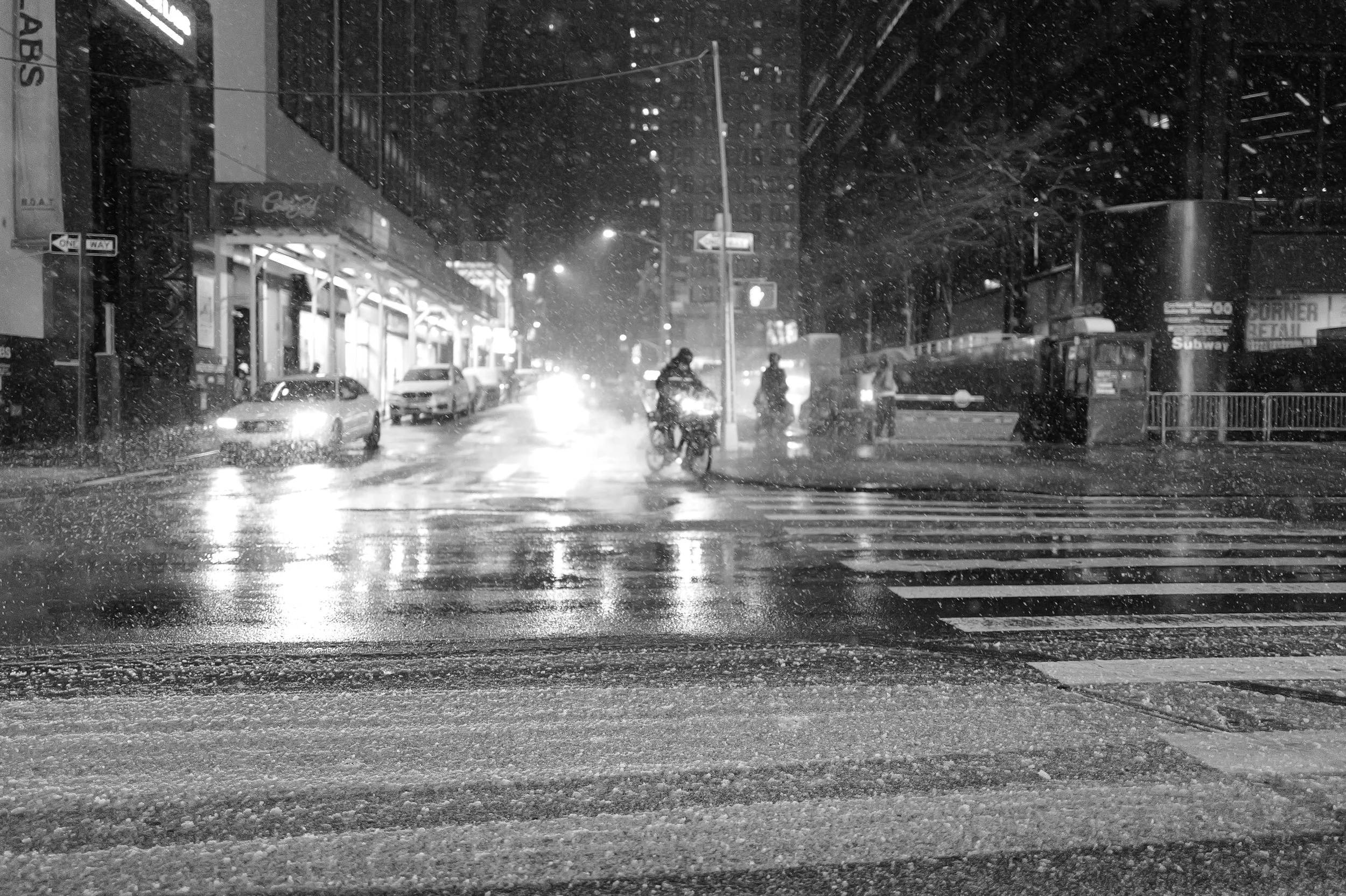 Une rue urbaine la nuit sous la pluie, avec des voitures, un bus et des piétons, certains sur un vélo, dans un environnement animé
