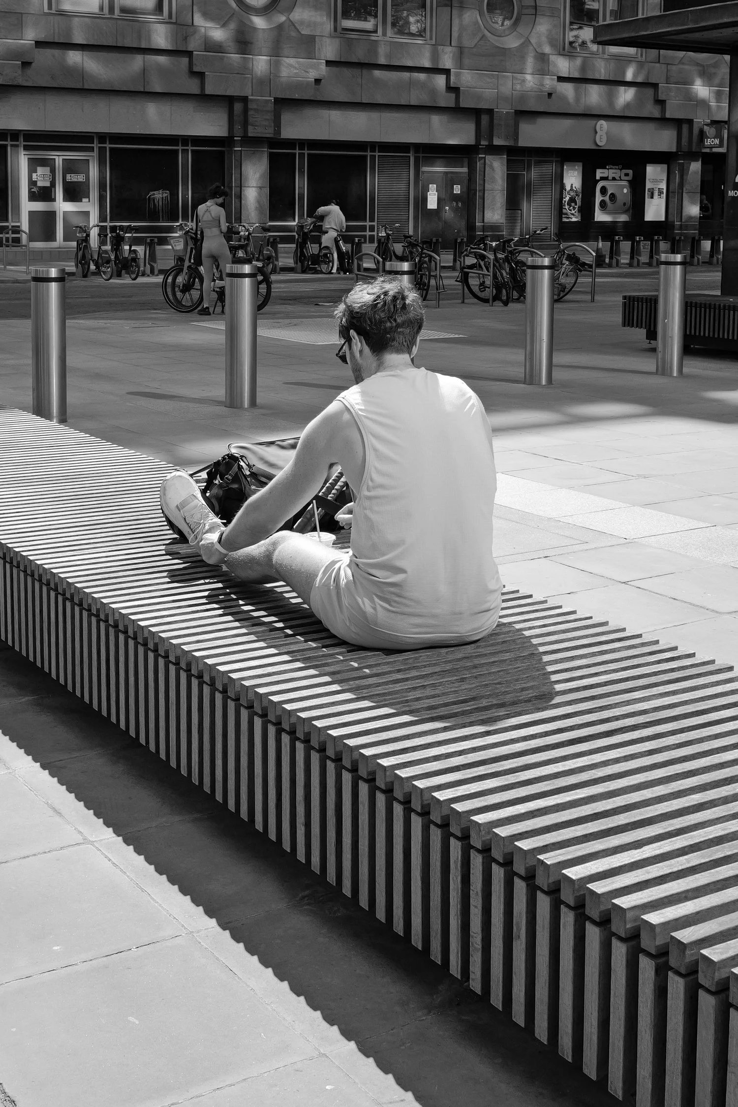 Un homme assis sur un banc en bois, portant un t-shirt sans manches et des shorts, dans une zone urbaine bordée de cabines de vélos et de bâtiments modernes