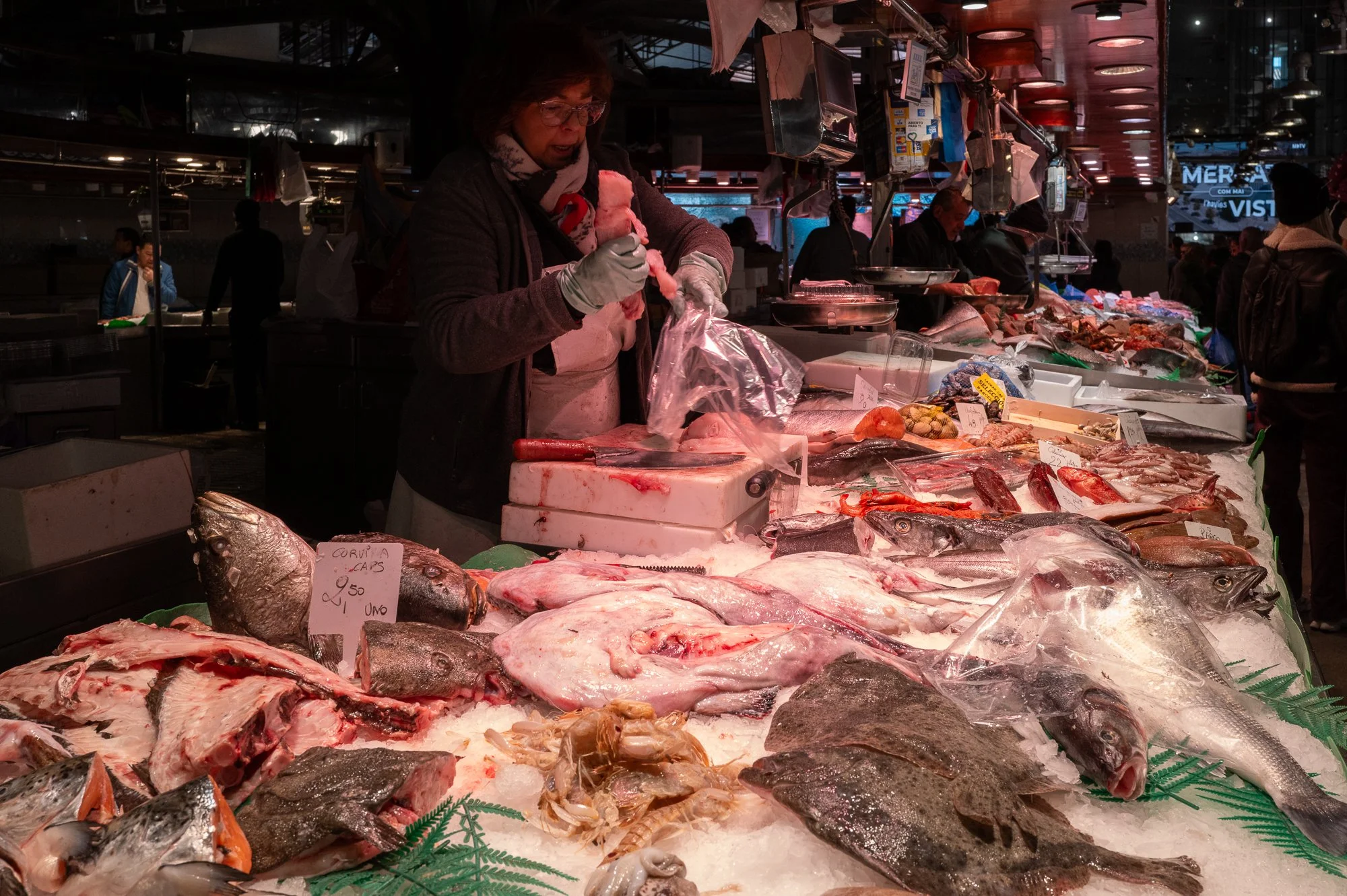 Vendeur de poisson dans un marché, devant une étalage de poissons entiers et coupes, à l'intérieur ou en extérieur, avec plusieurs personnes en arrière-plan.