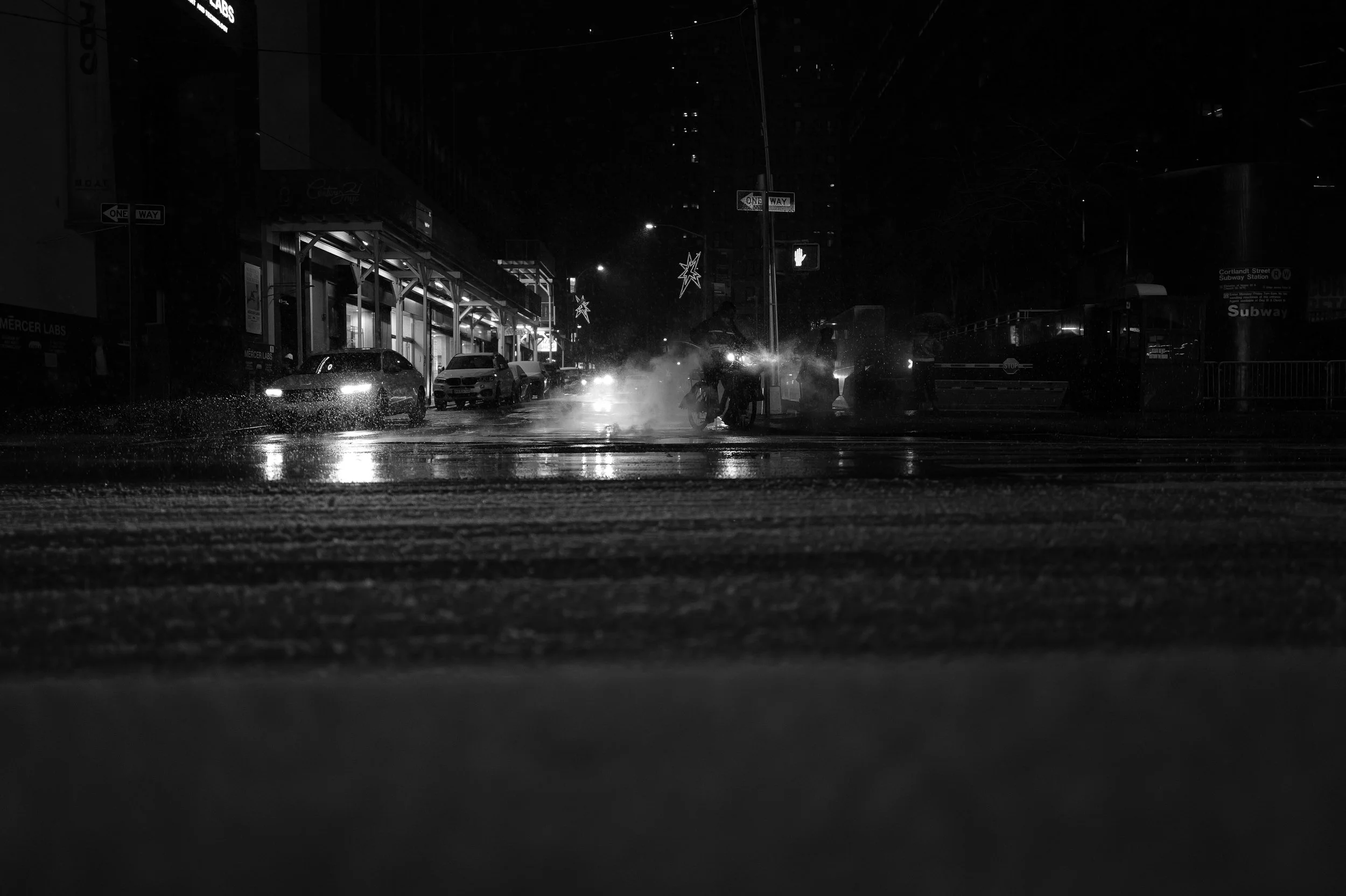 Une rue de nuit avec des voitures stationnées, de la vapeur ou de la pluie, des décorations lumineuses et une station de métro sous la pluie, dans une ambiance sombre.