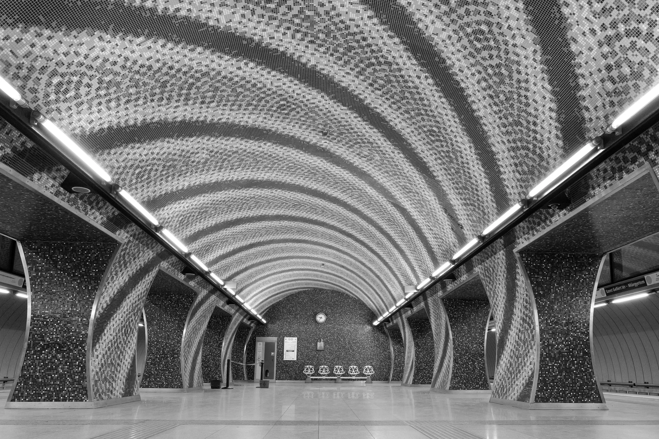 Hall d'entrée d'une station de métro à plafond incurvé décoré de mosaïques colorées, avec des lampes lumineuses, une horloge, un panneau d'information et des sièges.