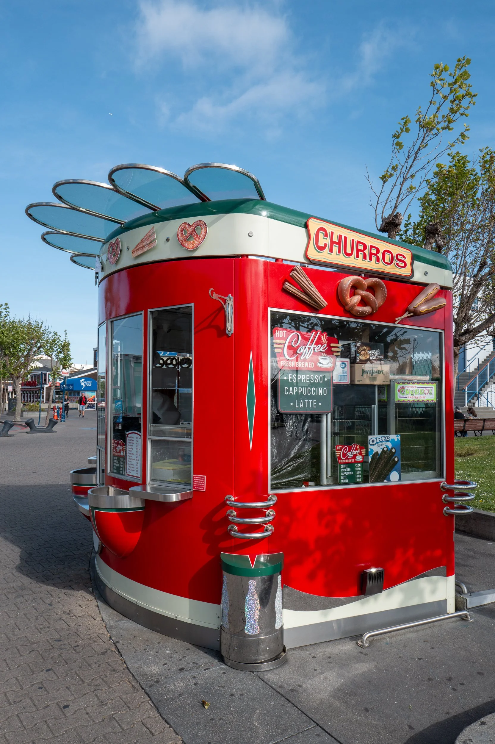 Kiosque à churros rouge avec décor de pretzels, hot dog, et cuillères à la vanille, vendant principalement des churros, café, et autres boissons.
