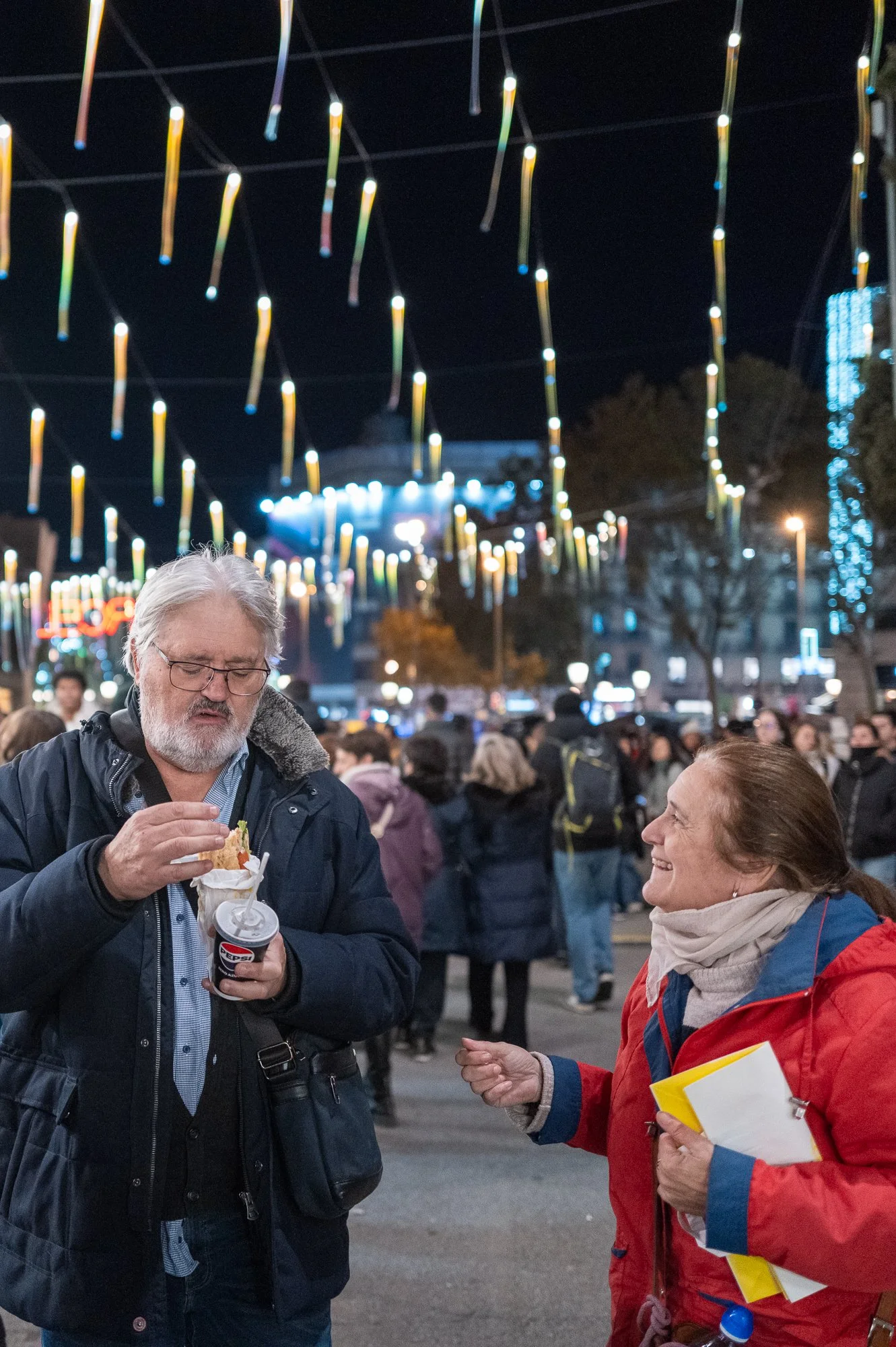 Deux personnes parlant lors d'un événement nocturne en plein air, avec des décorations lumineuses suspendues au-dessus et une foule de personnes en arrière-plan.