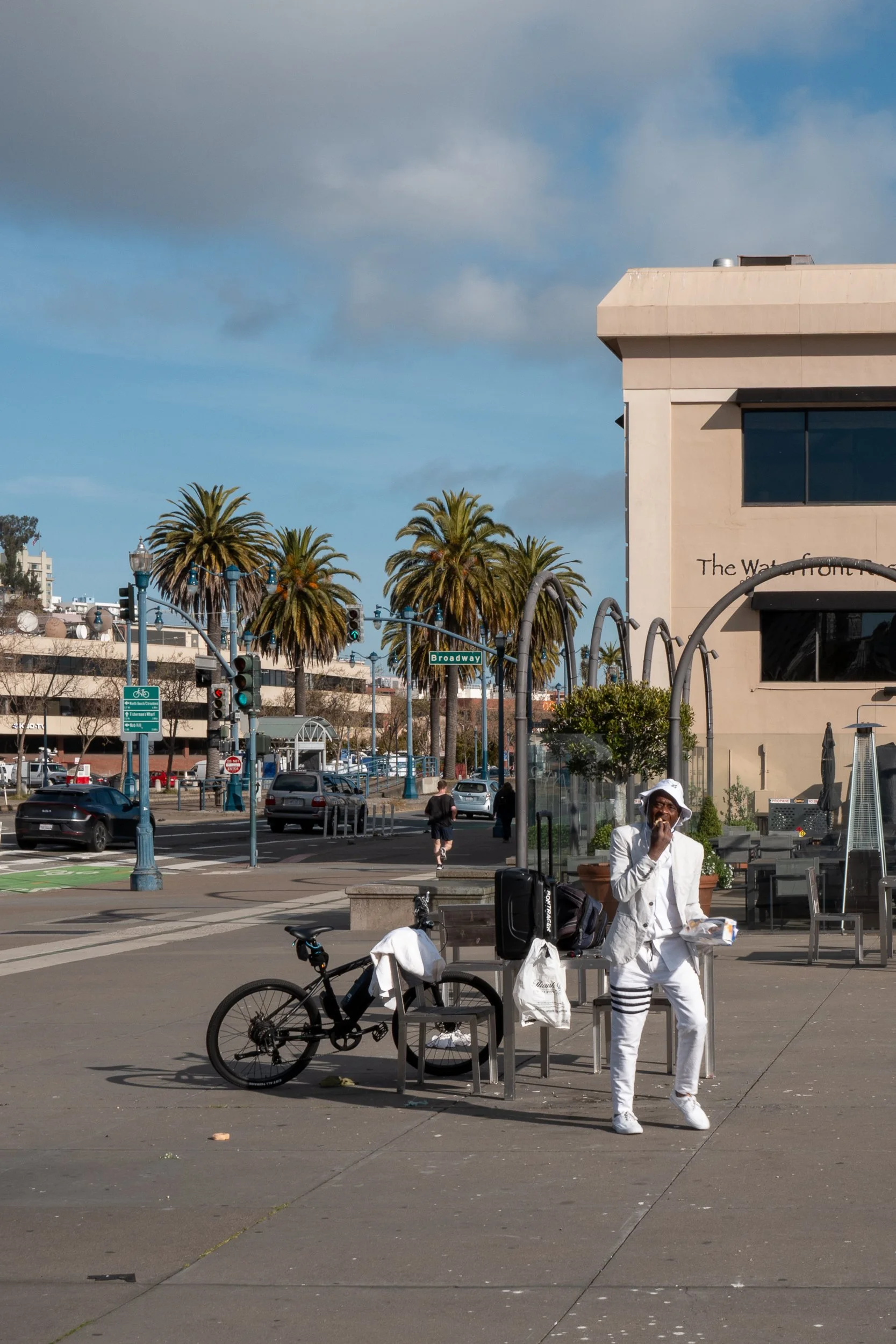 Une personne habillée en blanc avec un chapeau blanc, assise sur un banc, en train de manger ou de boire, avec un vélo à côté, dans une zone urbaine ensoleillée avec des palmiers et des bâtiments modernes en arrière-plan.