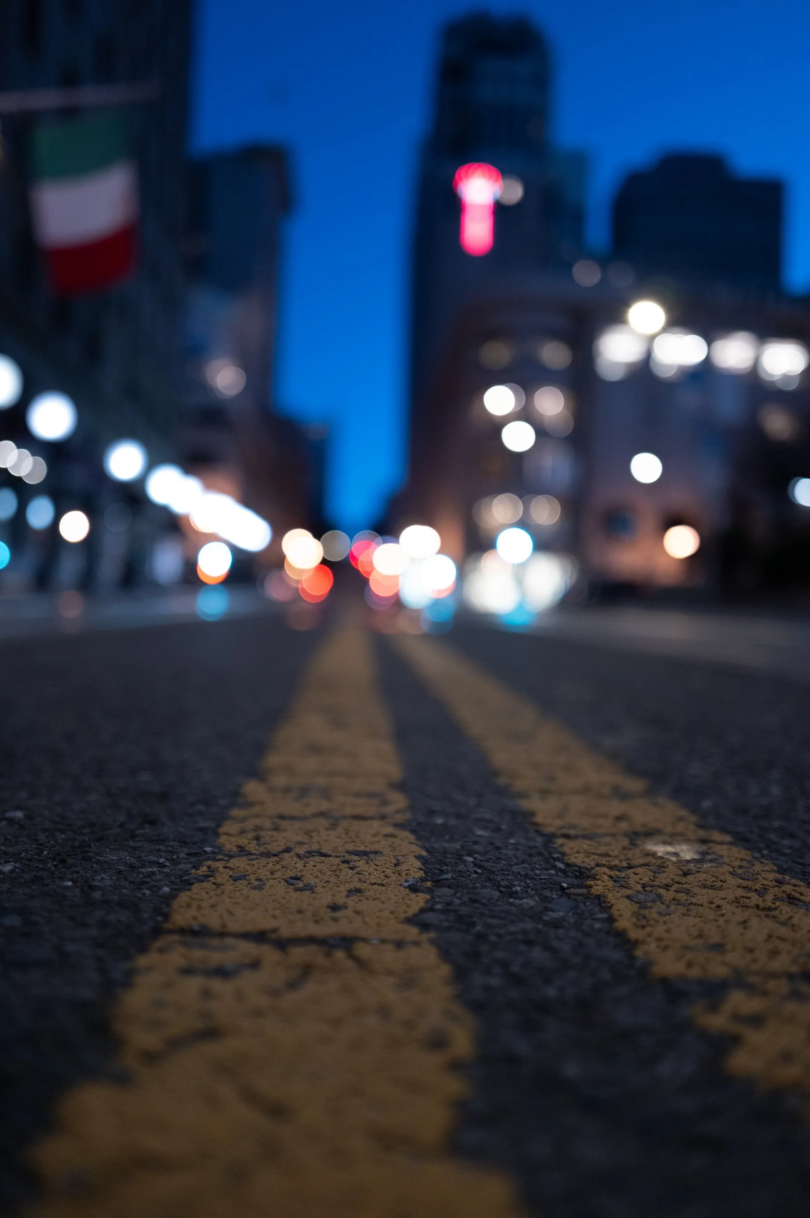 Vue en plongée d'une rue urbaine la nuit, avec une double ligne jaune centrale, floue en arrière-plan avec des bâtiments, des lumières de ville et un ciel bleu foncé.