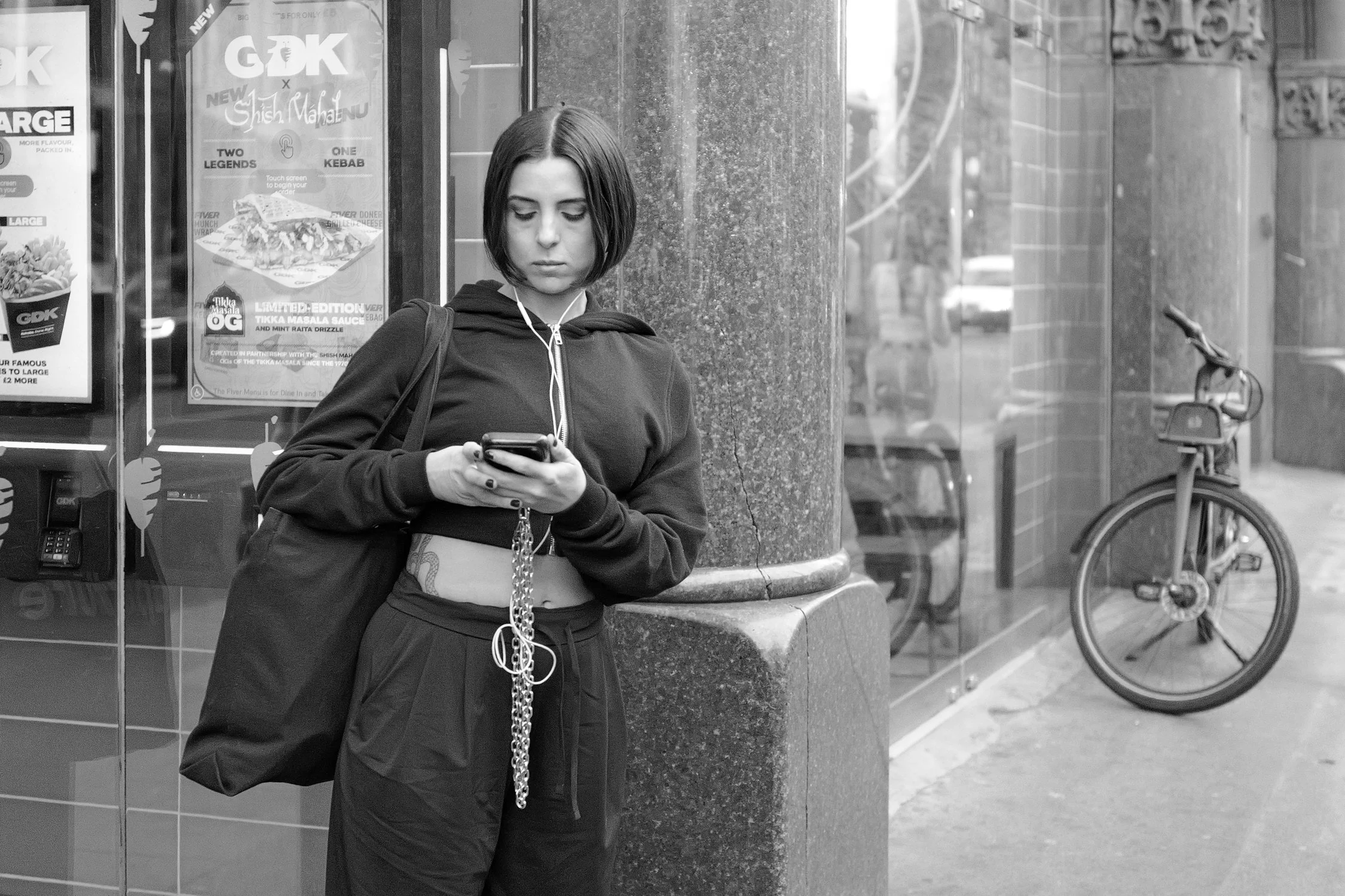 Jeune femme avec des cheveux courts, portant une veste à capuche noire, tenant un téléphone portable, debout à côté d'une colonne en pierre dans la rue. Elle a des écouteurs connectés et un sac noir sur l'épaule.